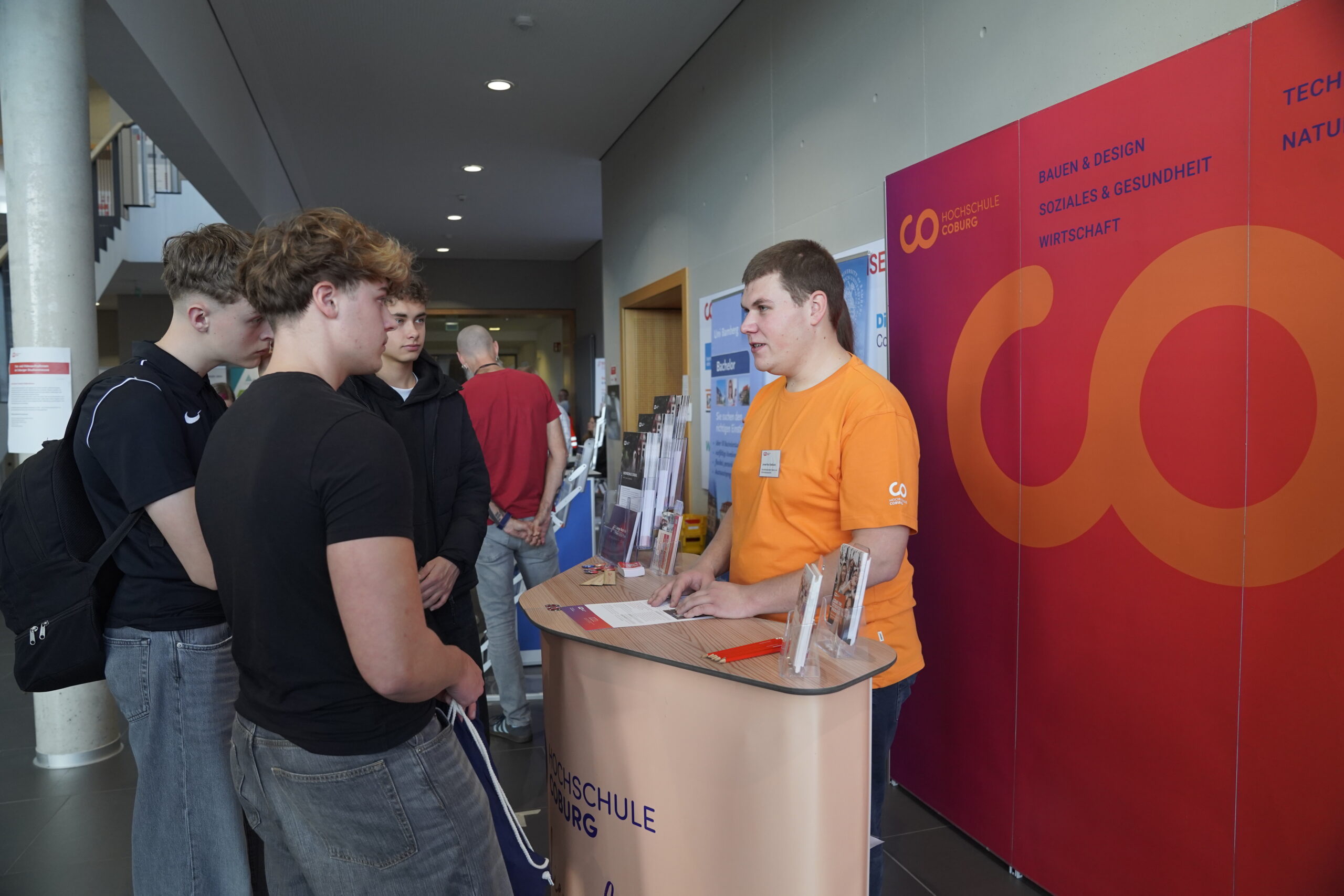Ein junger Mann in einem orangefarbenen Hemd steht hinter einem Informationsschalter und spricht mit drei Studenten in Freizeitkleidung auf der Coburger Studienmesse 2025. Im Hintergrund sind Informationsbanner zu sehen, die Orientierung für die Zukunft bieten. at Hochschule Coburg