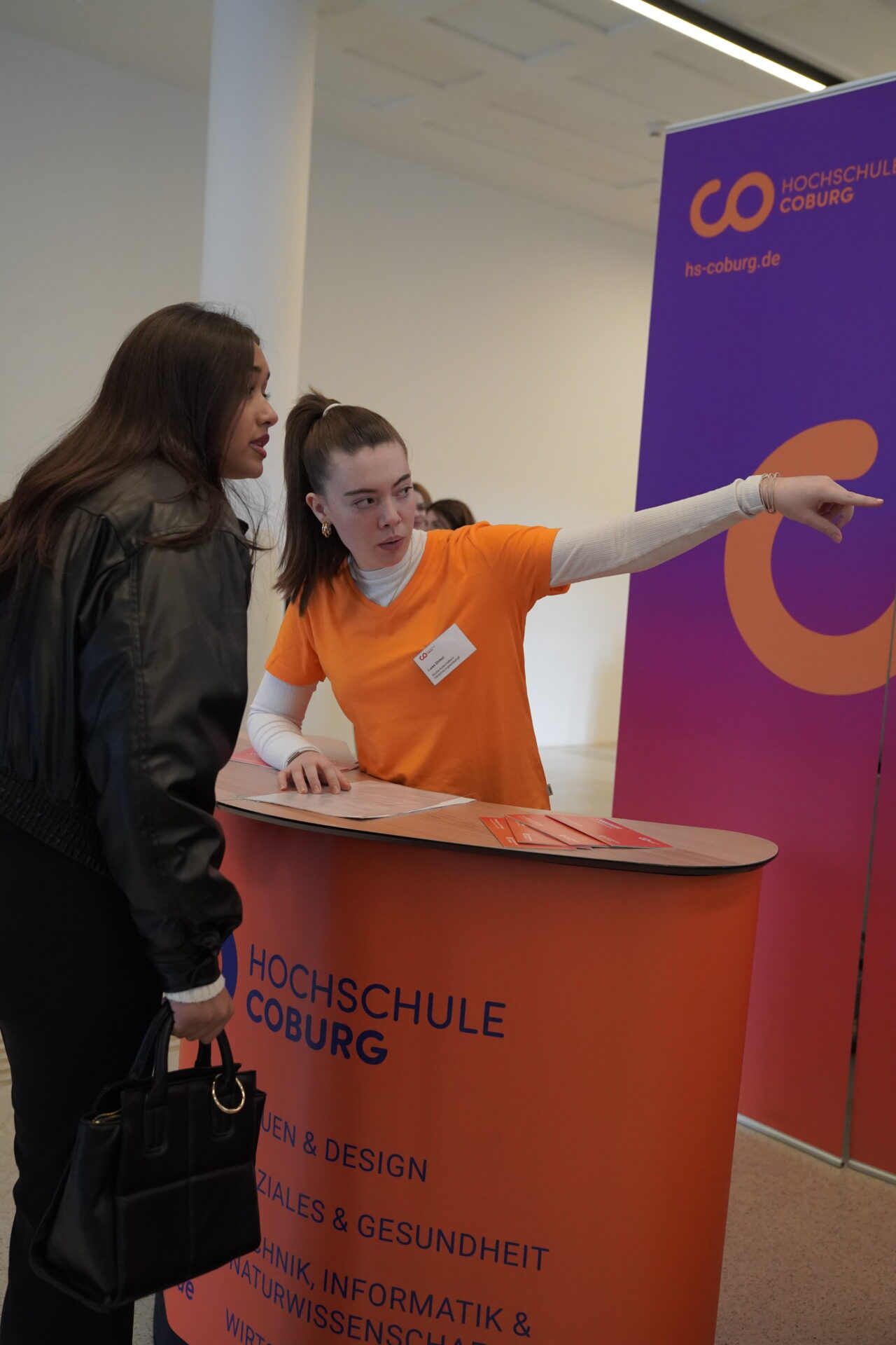 Eine Frau in einem orangefarbenen Hemd steht hinter einem Infotisch mit der Aufschrift "Hochschule Coburg" und zeigt auf den Tisch, während sie über die Orientierung Zukunft auf der Coburger Studienmesse 2025 informiert, während eine andere Frau in einer schwarzen Jacke zuhört. Im Hintergrund ist ein Banner der Hochschule Coburg zu sehen. at Hochschule Coburg