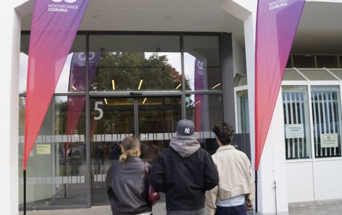 Drei Personen gehen auf den gläsernen Eingang eines Gebäudes mit der Aufschrift "5" zu, flankiert von hohen lila-roten Veranstaltungsfahnen mit der Aufschrift "Coding Hall" auf der Coburger Studienmesse 2025 - einer Veranstaltung zur Orientierung für die Zukunft auf einem gepflasterten Gehweg. at Hochschule Coburg