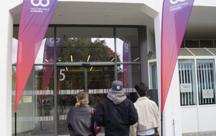 Drei Personen gehen auf den gläsernen Eingang eines Gebäudes mit der Aufschrift "5" zu, flankiert von hohen lila-roten Veranstaltungsfahnen mit der Aufschrift "Coding Hall" auf der Coburger Studienmesse 2025 - einer Veranstaltung zur Orientierung für die Zukunft auf einem gepflasterten Gehweg. at Hochschule Coburg