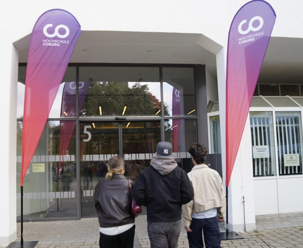 Drei Personen gehen auf den gläsernen Eingang eines Gebäudes mit der Aufschrift "5" zu, flankiert von hohen lila-roten Veranstaltungsfahnen mit der Aufschrift "Coding Hall" auf der Coburger Studienmesse 2025 - einer Veranstaltung zur Orientierung für die Zukunft auf einem gepflasterten Gehweg. at Hochschule Coburg