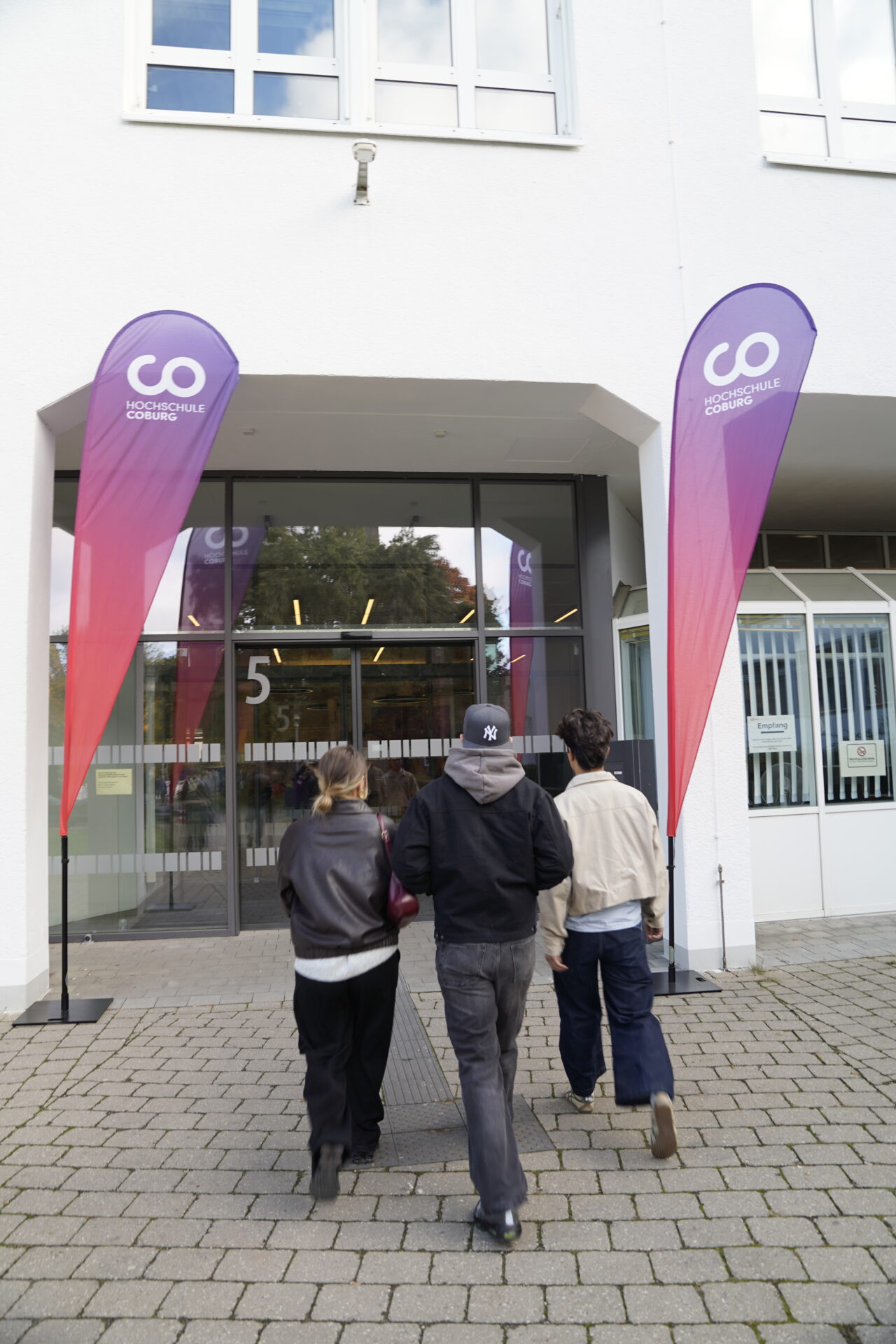 Drei Personen gehen auf den gläsernen Eingang eines Gebäudes mit der Aufschrift "5" zu, flankiert von hohen lila-roten Veranstaltungsfahnen mit der Aufschrift "Coding Hall" auf der Coburger Studienmesse 2025 - einer Veranstaltung zur Orientierung für die Zukunft auf einem gepflasterten Gehweg. at Hochschule Coburg