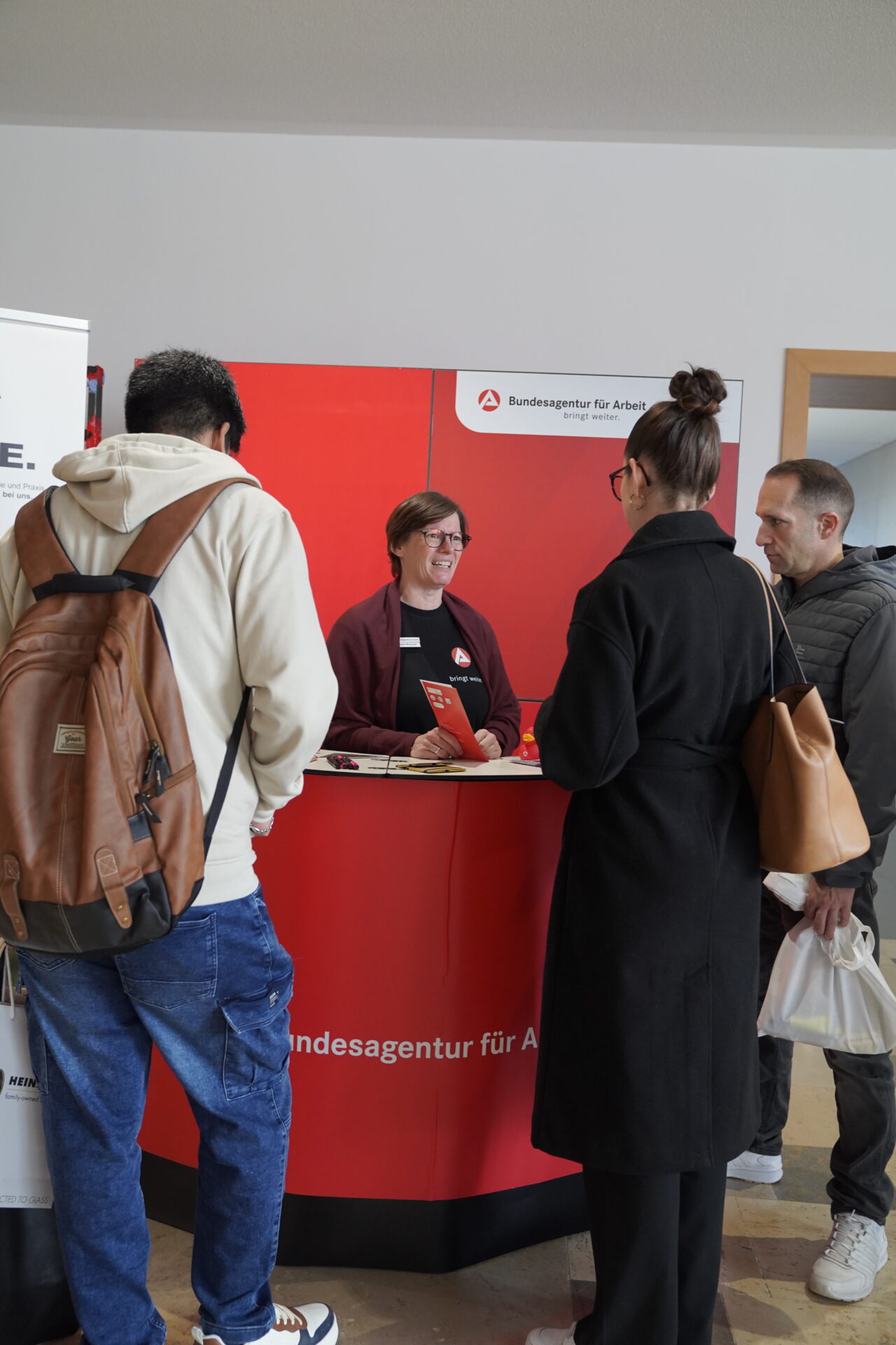 Drei Personen stehen während der Coburger Studienmesse 2025 an einem roten Stand der Bundesagentur für Arbeit, wo eine lächelnde Frau hinter dem Tresen Orientierung für die Zukunft bietet, indem sie hilfreiche Informationen an die Besucherinnen und Besucher weitergibt. at Hochschule Coburg