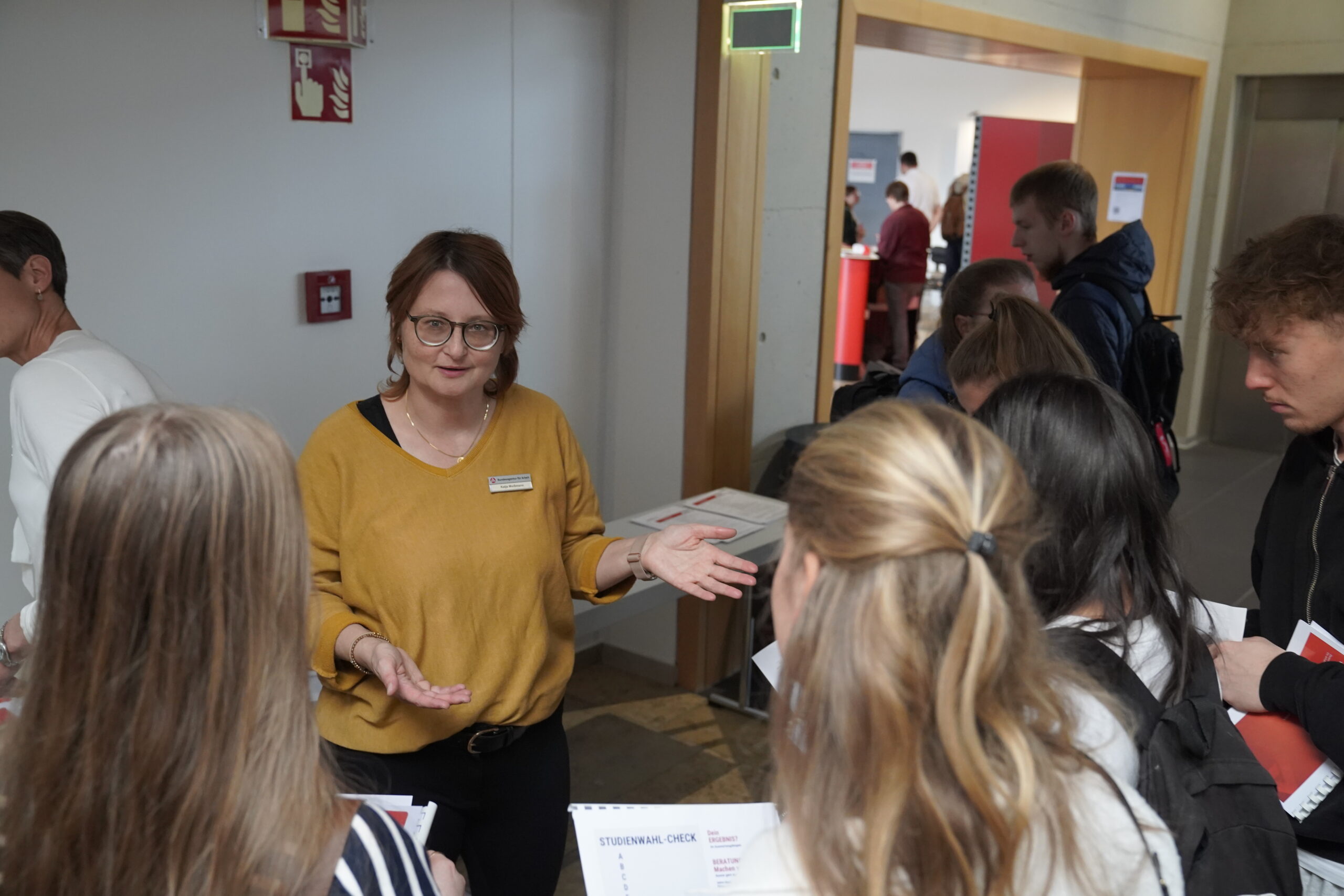 Eine Frau mit Brille und gelbem Oberteil spricht in einem Flur der Coburger Studienmesse 2025 zu einer Gruppe von Menschen, die Papiere in der Hand halten, und bietet Orientierung für die Zukunft. Andere Personen sind im Hintergrund in der Nähe eines Durchgangs zu sehen. at Hochschule Coburg