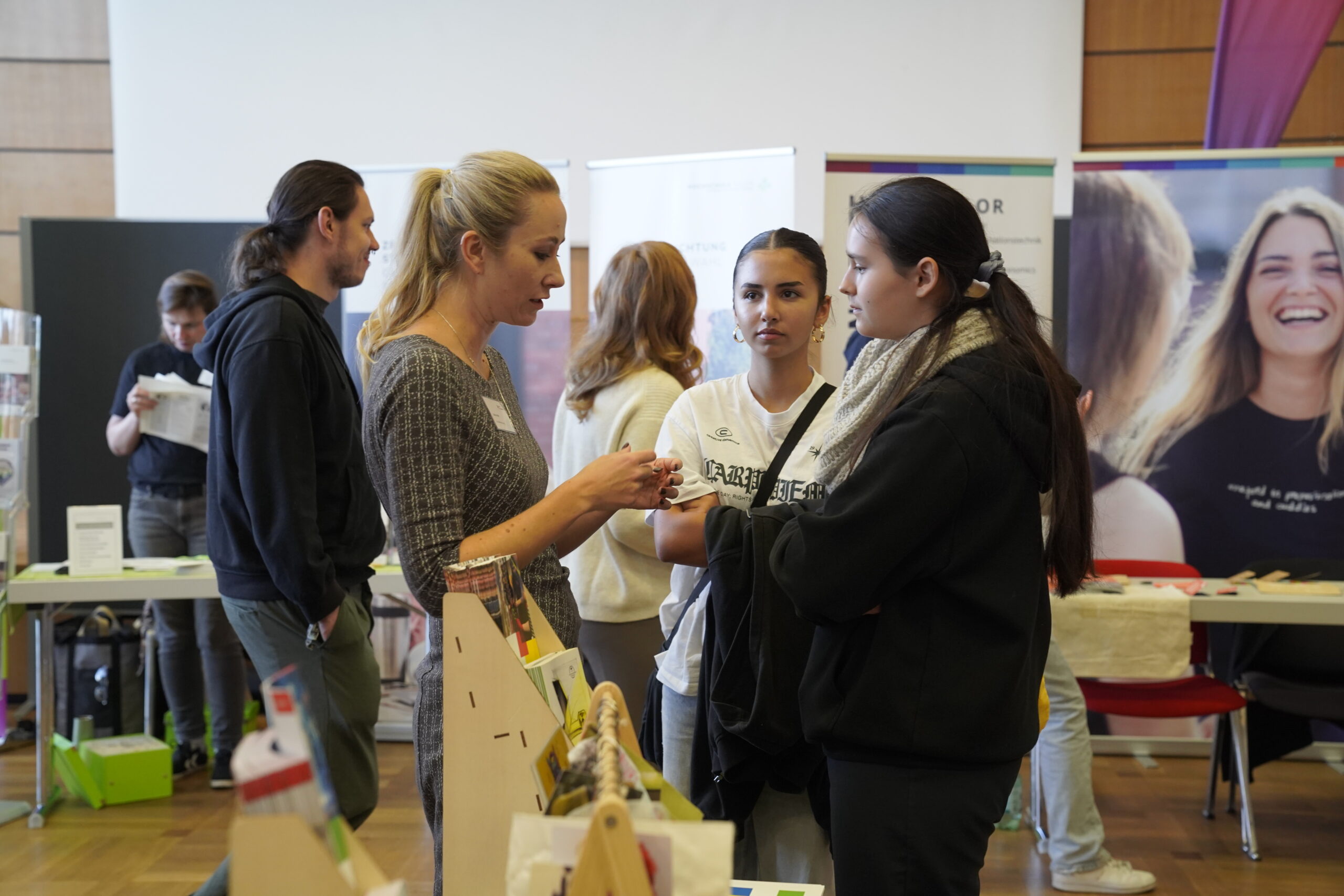 Eine Gruppe von Menschen im Gespräch bei einer Indoor-Veranstaltung mit Informationsständen und Displays im Hintergrund. Auf der Coburger Studienmesse 2025 hören zwei junge Frauen einer älteren Frau zu, die Orientierung für die Zukunft anbietet. at Hochschule Coburg