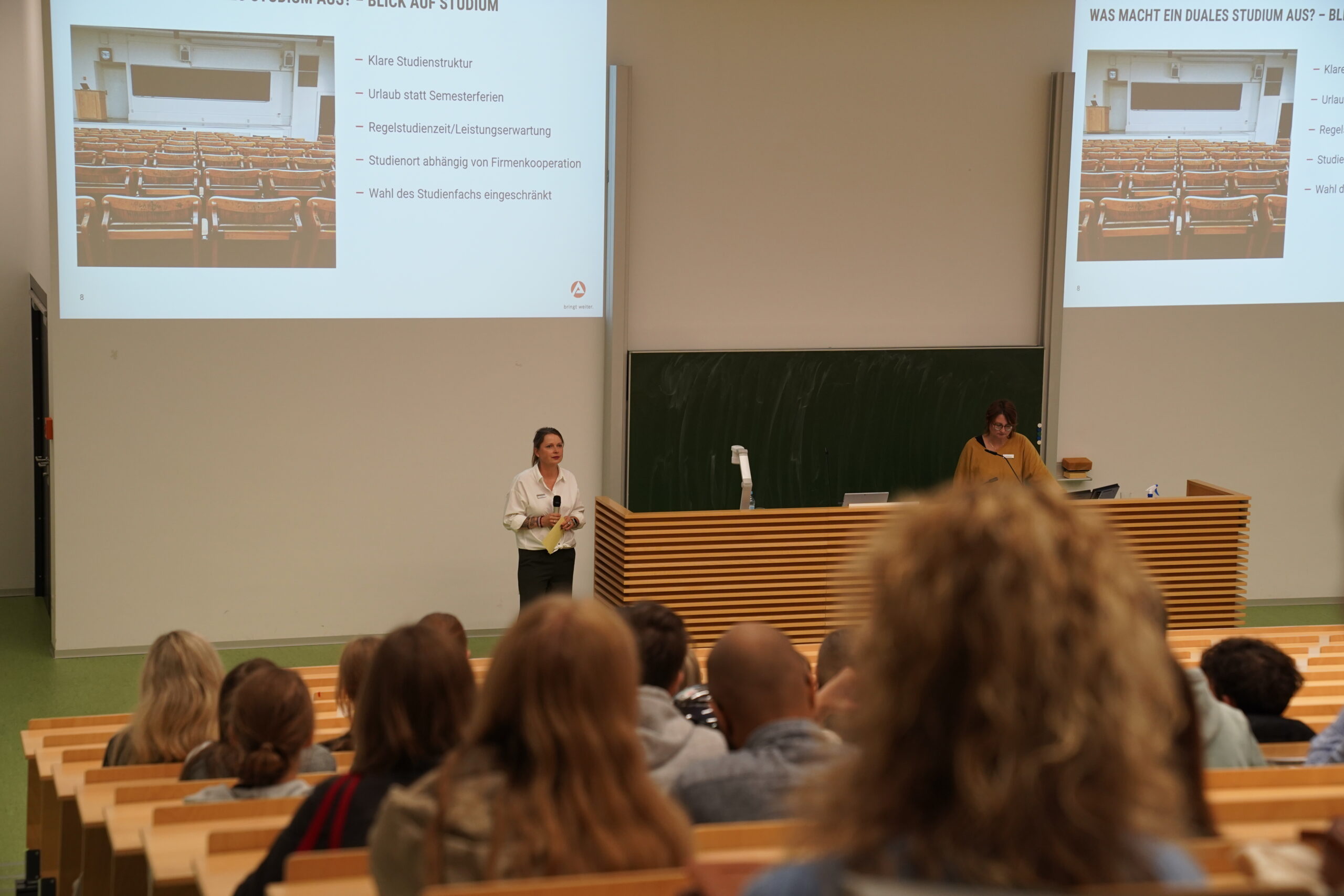 Ein Klassenzimmer mit Schülern, die zwei Referenten gegenübersitzen, von denen einer spricht und einer an einem Pult steht. An der Wand über ihnen werden Dias über das Studium auf der Coburger Studienmesse 2025 projiziert. at Hochschule Coburg