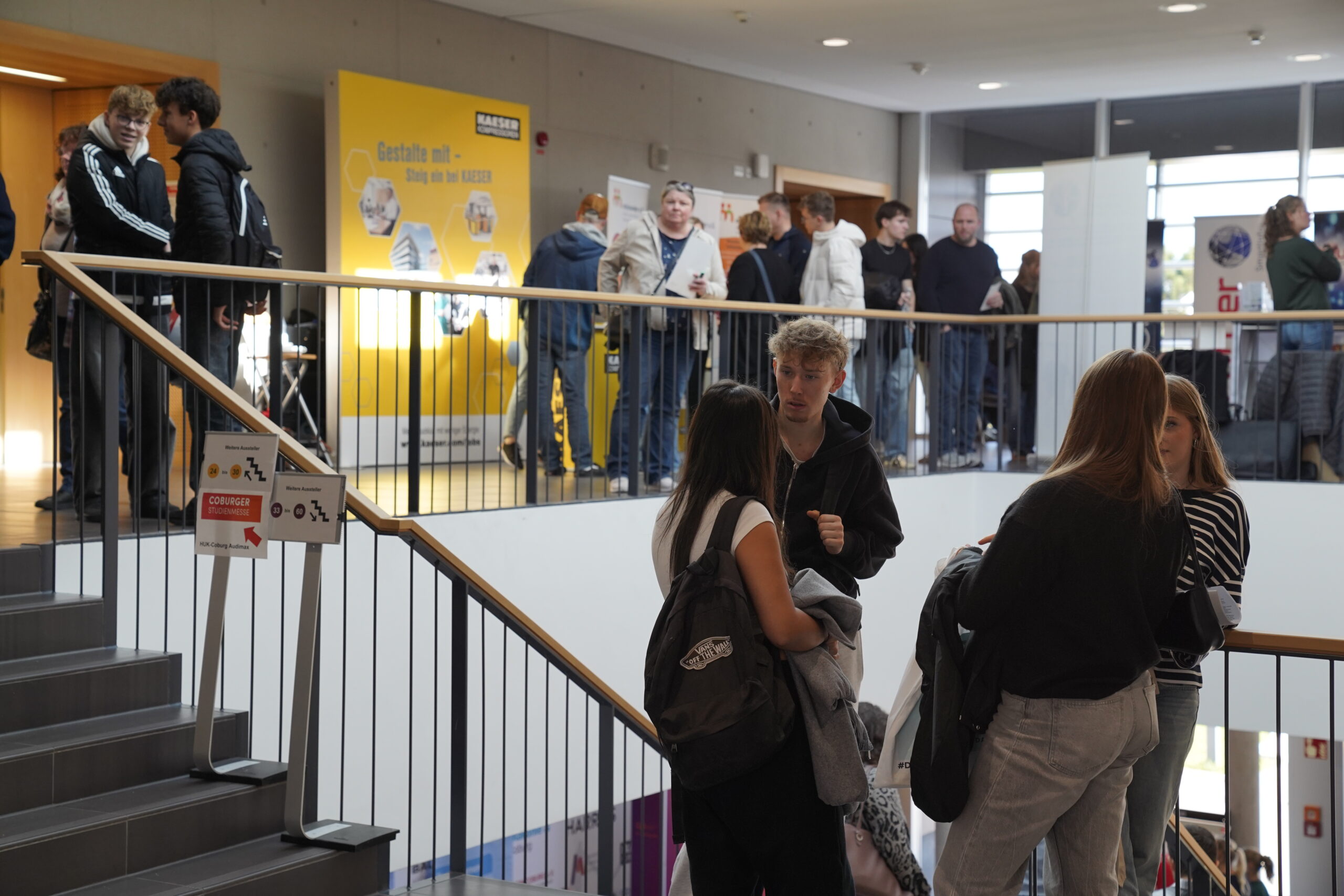 Eine Gruppe von Menschen steht und unterhält sich auf einer Treppe und einem offenen Treppenabsatz in einem modernen Gebäude während der Coburger Studienmesse 2025. Andere reihen sich in einem Flur auf, wo leuchtend gelbe Orientierungsplakate an der Wand zu sehen sind. at Hochschule Coburg