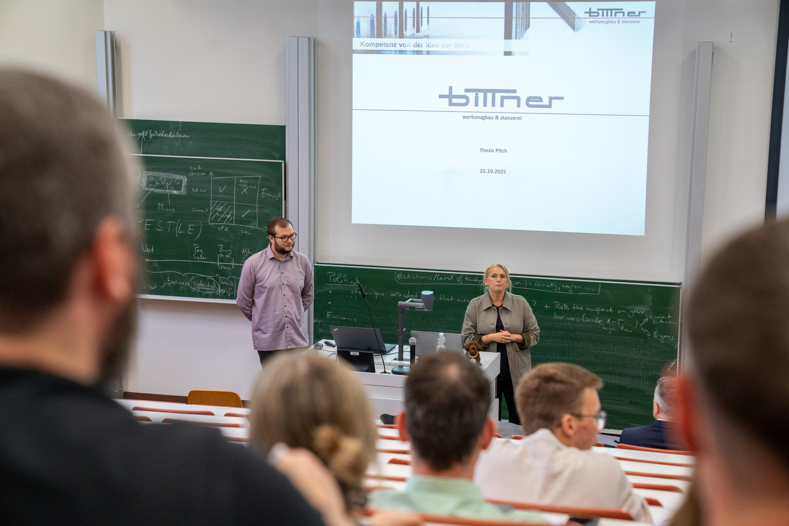 Eine Frau und ein Mann stehen vorne in einem Hörsaal und sprechen zu einem Publikum über Themen und Abschlussarbeiten. Hinter ihnen stehen beschriftete Kreidetafeln und eine große Präsentationswand mit dem Titel "BITNER. at Hochschule Coburg