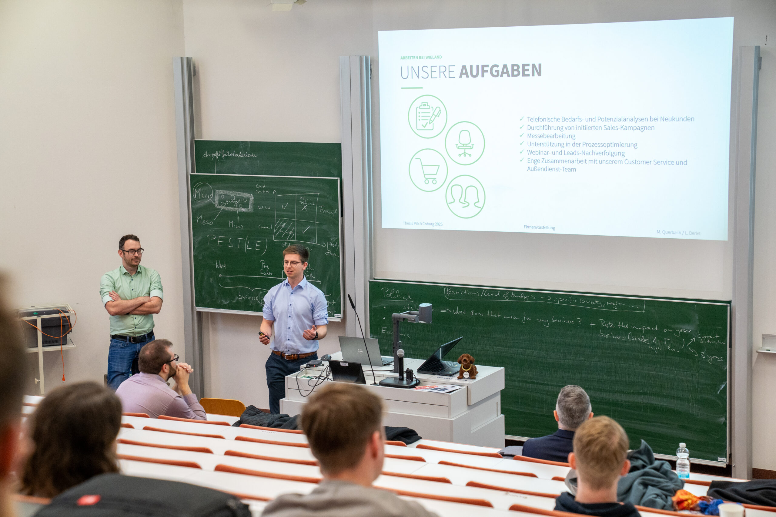 Mehrere Personen sitzen in einem Vortragssaal, während zwei Männer an der Stirnseite neben einem Projektor stehen und "UNSERE AUFGABEN" präsentieren. Im Hintergrund sind Kreidetafeln mit Schrift und Themen zu Unternehmen und Abschlussarbeiten zu sehen. at Hochschule Coburg