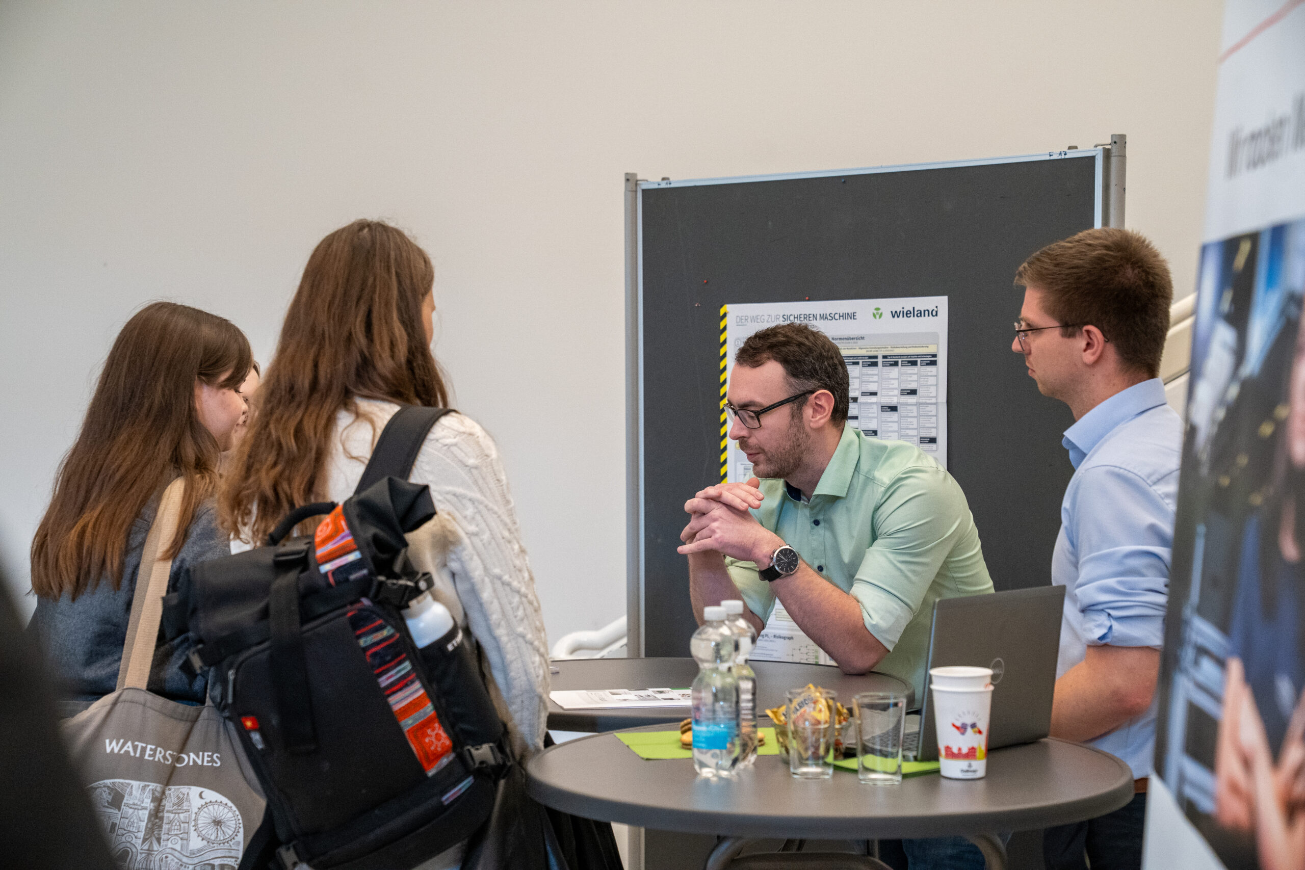 Drei junge Frauen unterhalten sich mit zwei Männern an einem Stand, der wie eine Karrieremesse aussieht. Sie diskutieren über Abschlussarbeiten und Themen, auf dem Tisch liegen Firmenflyer, Getränke und ein Laptop. at Hochschule Coburg