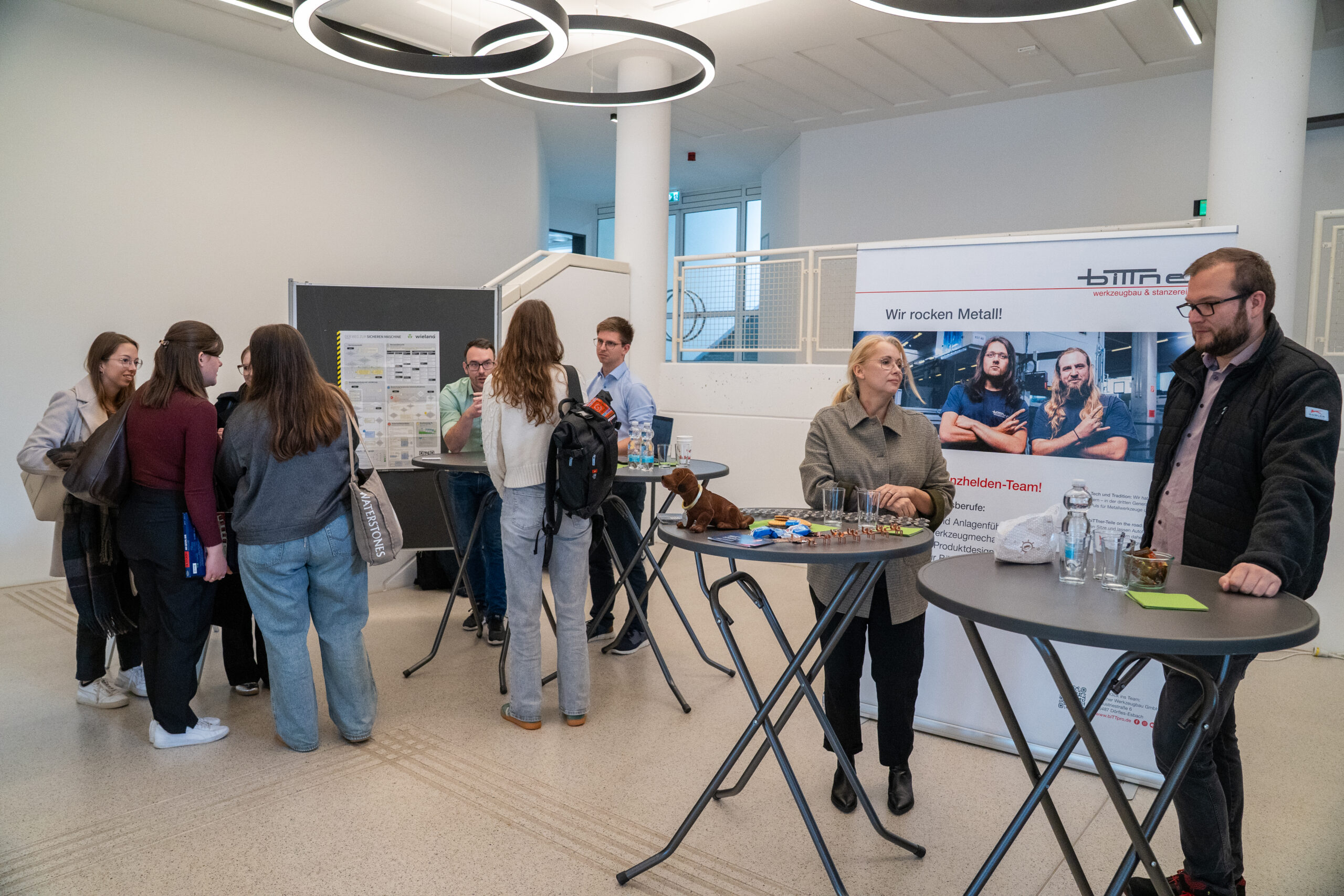 Eine Gruppe von Menschen steht und unterhält sich um hohe Tische bei einer Indoor-Unternehmensveranstaltung mit Plakaten und Werbematerialien. Zwei Personen stehen an einem Stand mit einem Schild "Wir rocken Metall!" und Markenartikeln auf dem Tisch. at Hochschule Coburg