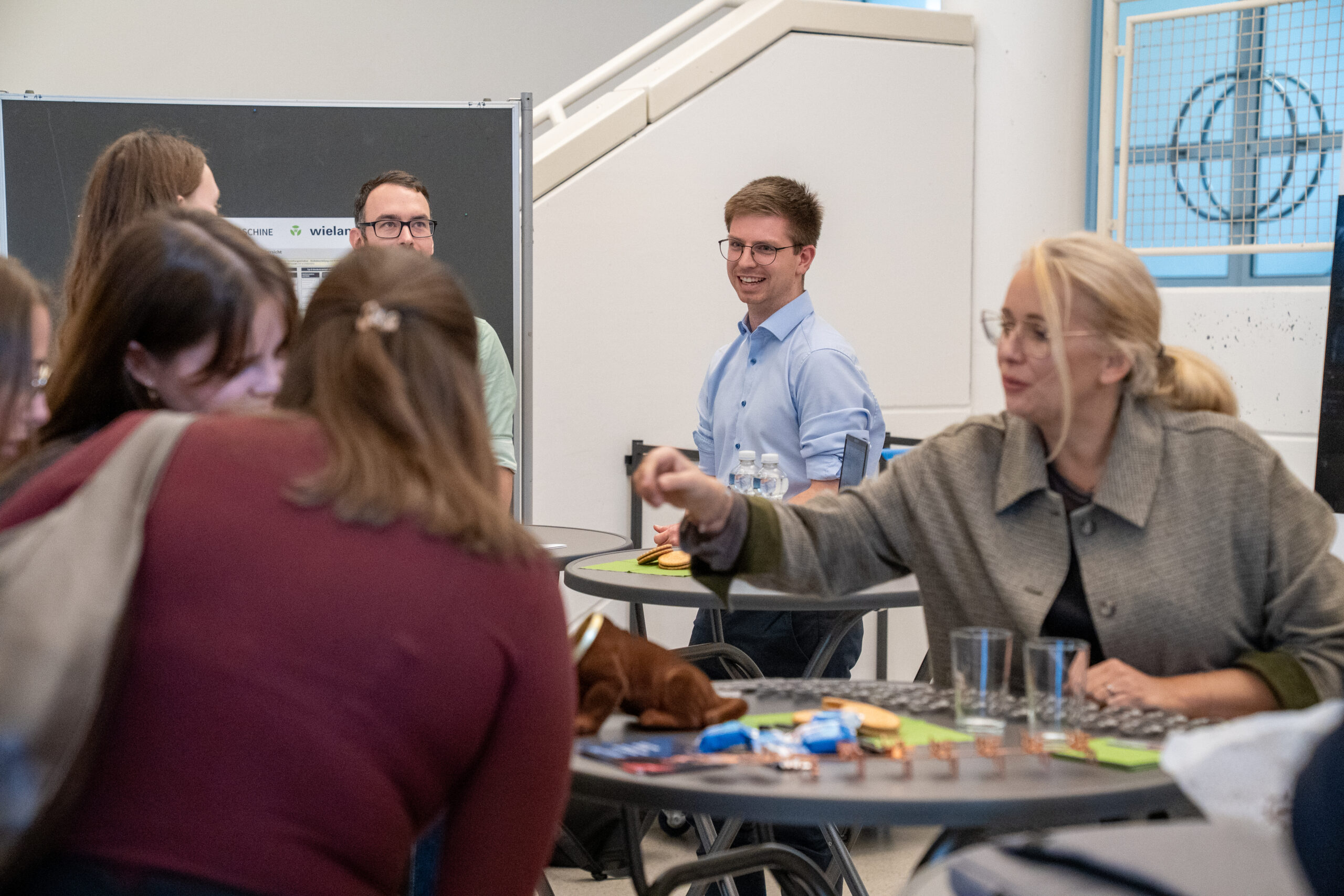 Mehrere Menschen sitzen und stehen um kleine Tische herum und unterhalten sich über Abschlussarbeiten und Themen. Eine Frau greift nach Gegenständen auf dem Tisch, während andere sich in einem hellen, modernen Innenraum unterhalten und lächeln. at Hochschule Coburg