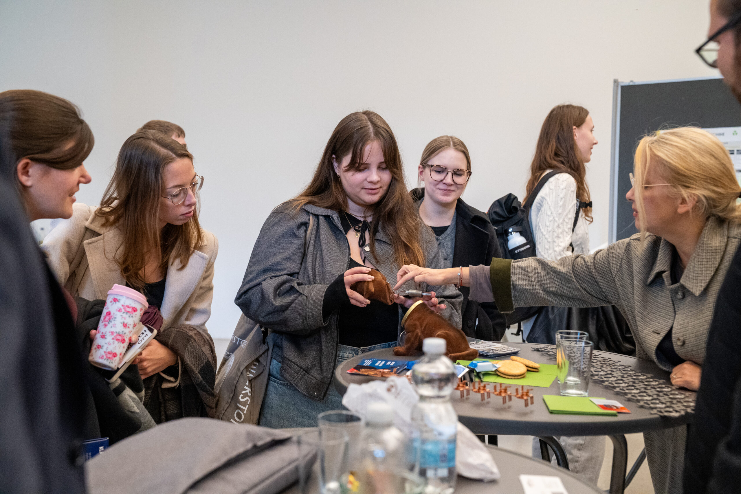 Eine Gruppe von Menschen steht um einen hohen Tisch herum, unterhält sich und lacht. Eine Frau greift nach einem Gegenstand auf dem Tisch, der mit Snacks und Getränken gedeckt ist, während sie in einer lockeren, freundlichen Atmosphäre über Themen im Zusammenhang mit Unternehmen diskutieren. at Hochschule Coburg