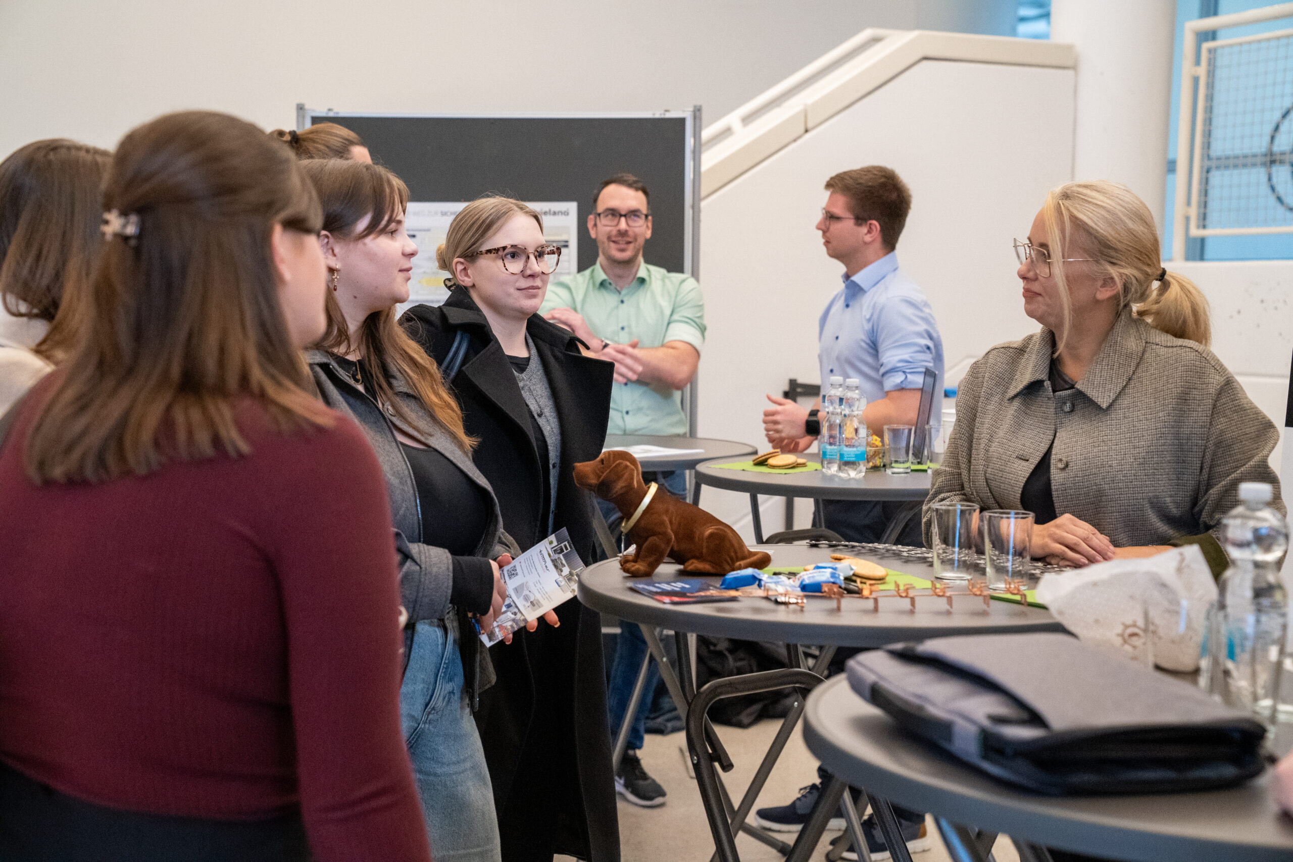 Eine Gruppe von Menschen steht bei einer Veranstaltung in einer Halle um Stehtische herum und diskutiert mit Vertretern von Unternehmen über Themen für Abschlussarbeiten. Einige halten Getränke in der Hand, während ein Mann beim Sprechen gestikuliert, und alle scheinen in ein Gespräch vertieft zu sein. at Hochschule Coburg