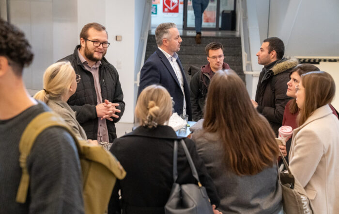 Eine Gruppe von Personen, einige stehend, einige einander zugewandt, unterhält sich in einem Innenraum in der Nähe einer Treppe. Die meisten tragen Jacken; eine Person hält eine Kaffeetasse in der Hand. Die Atmosphäre wirkt freundlich und professionell, mit Themen rund ums Unternehmen und Abschlussarbeiten im Gespräch. at Hochschule Coburg