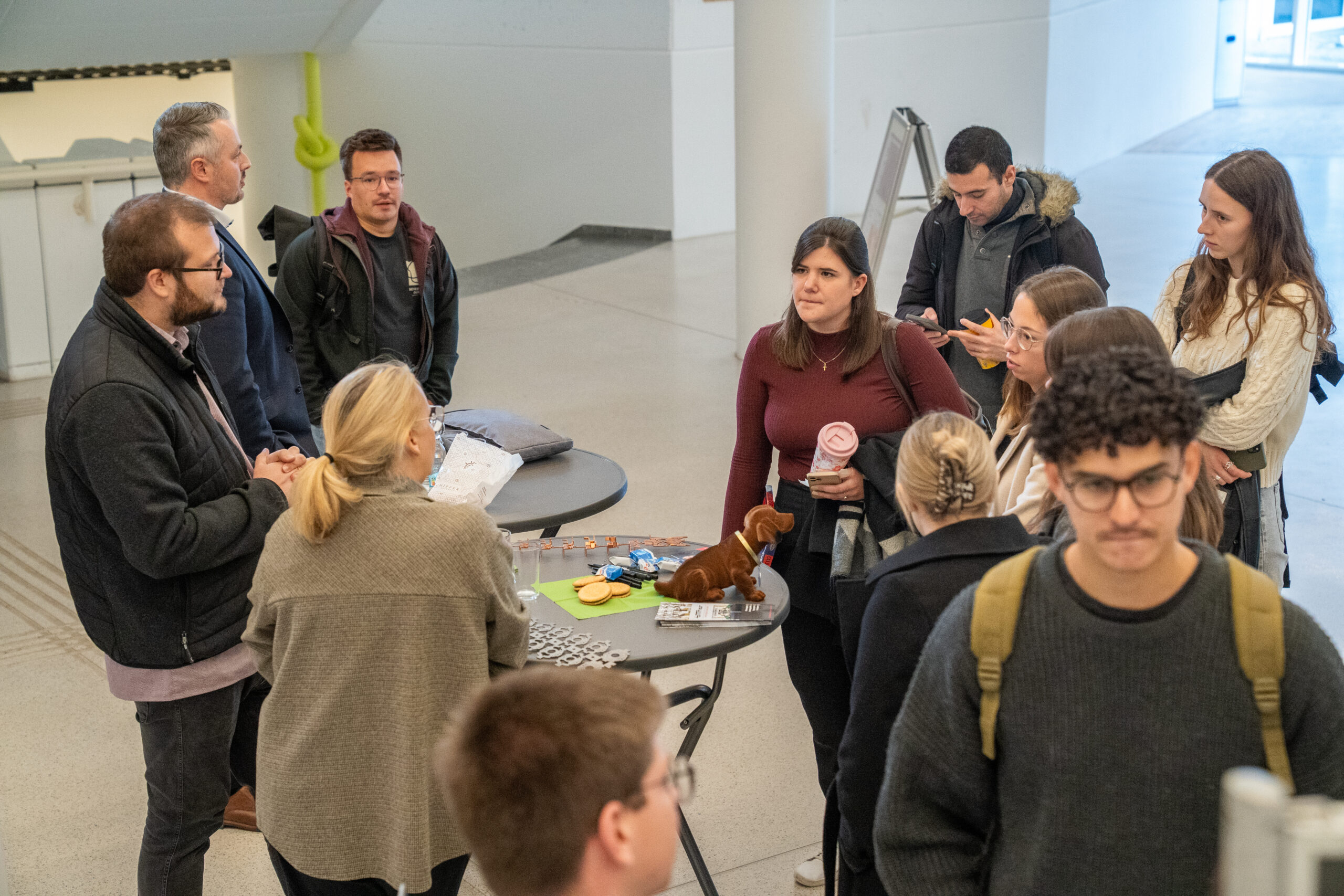 Eine Gruppe von Menschen sitzt an hohen Tischen in einem hellen Innenraum und unterhält sich über verschiedene Unternehmen und Themen. Einige halten Getränke in der Hand, und auf den Tischen liegen Gegenstände wie Knöpfe und ein kleiner Plüschhund, möglicherweise zur Diskussion über Abschlussarbeiten. at Hochschule Coburg