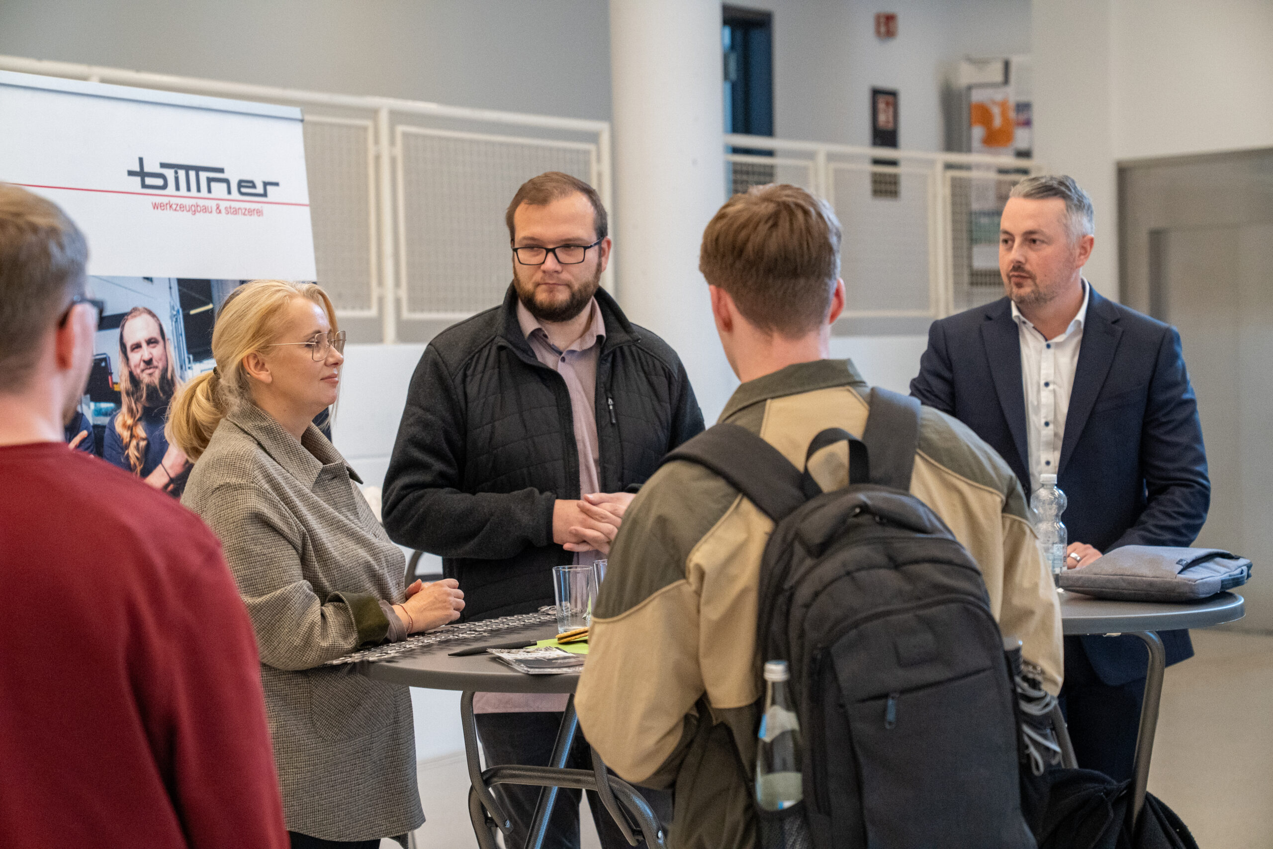 Vier Erwachsene unterhalten sich bei einer Indoor-Veranstaltung an Stehtischen, wobei Flyer und Wasserflaschen sichtbar sind. Im Hintergrund ist ein "Bittner"-Banner zu sehen, das auf Unternehmen und Themen für Abschlussarbeiten hinweist. Zwei Personen blicken von der Kamera weg. at Hochschule Coburg