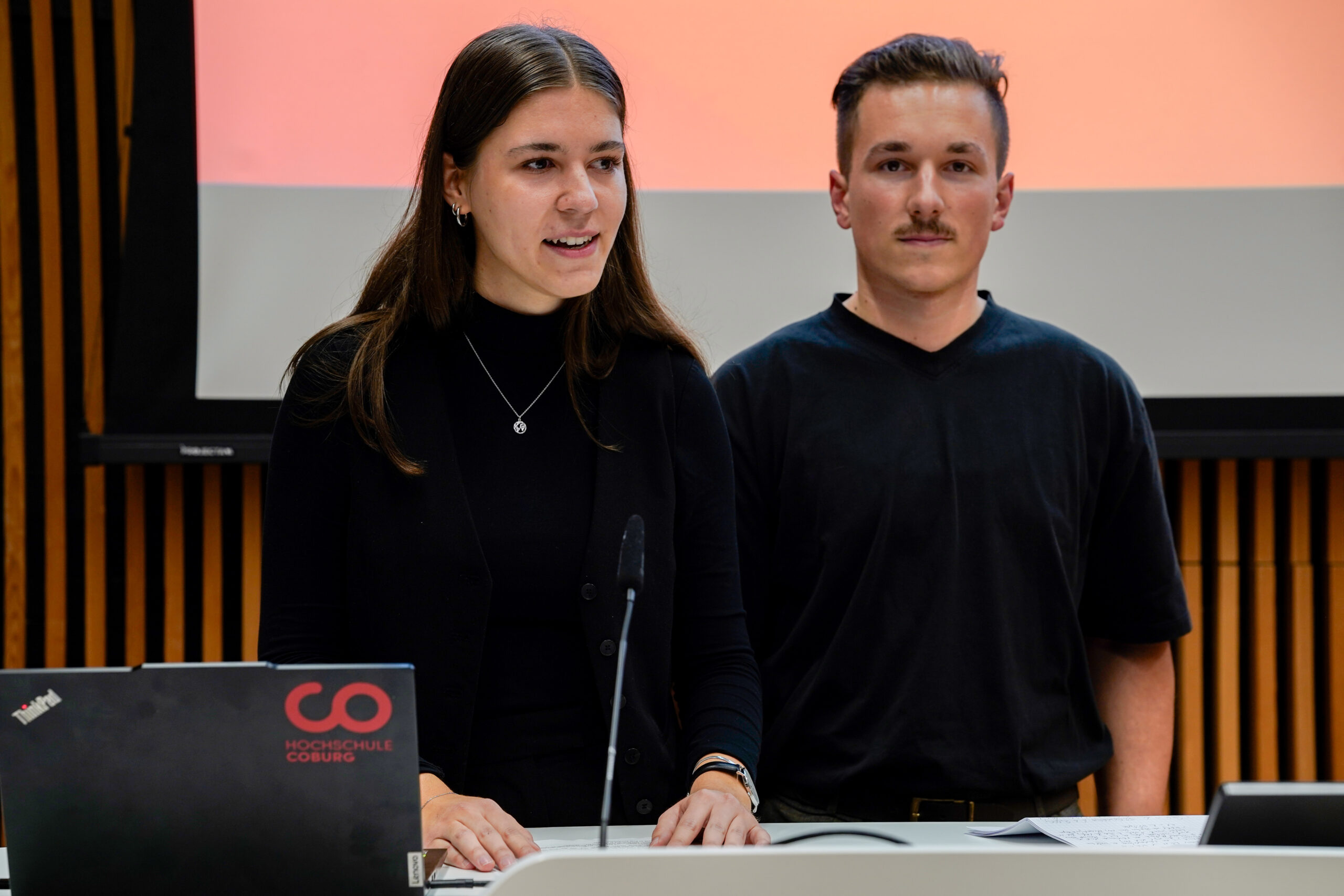 Zwei junge Erwachsene stehen an einem Podium und präsentieren vor einem Bildschirm. Die Frau spricht, während der Mann neben ihr steht. Beide sind schwarz gekleidet. Ein Laptop mit einem Aufkleber der Hochschule Coburg hebt ihre innovative Lehre hervor. at Hochschule Coburg