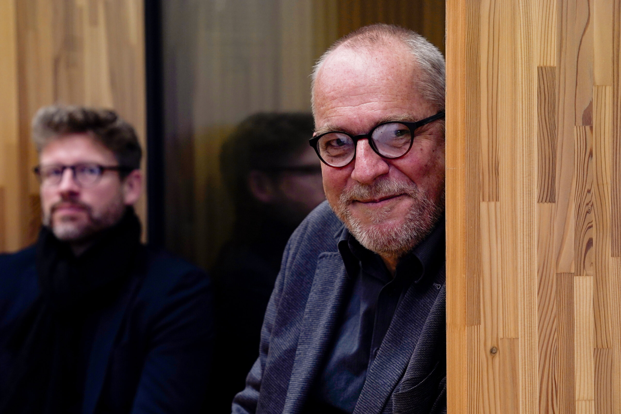 Zwei innovative Lehrende sitzen drinnen an einer Holzwand; der Mann im Vordergrund hat graues Haar, eine Brille, einen Bart und einen Anzug, während der Mann im Hintergrund, der unscharf ist, ebenfalls eine Brille und einen dunklen Schal trägt. at Hochschule Coburg