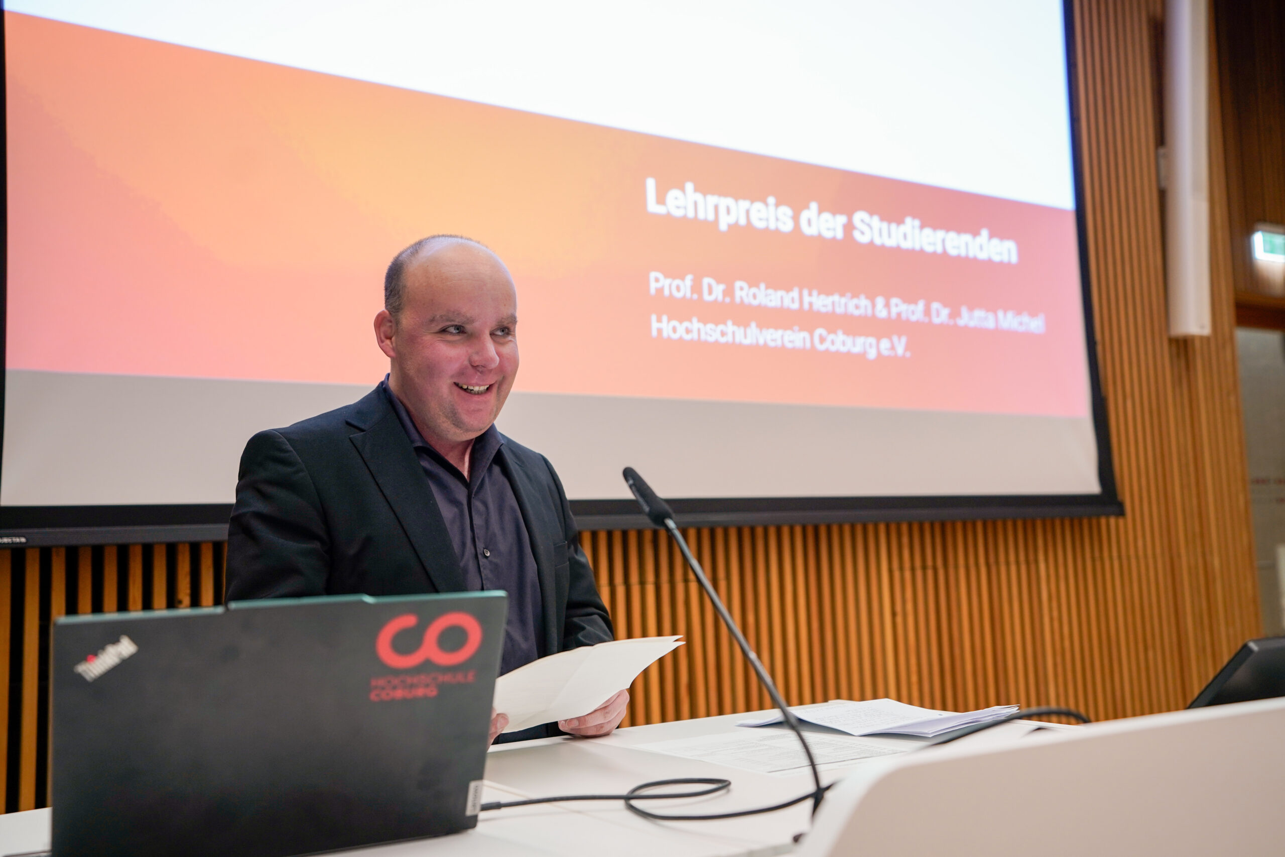 Ein Mann in einem dunklen Anzug steht an einem Podium, lächelt und hält Papiere in der Hand. Hinter ihm wird auf einem großen Bildschirm ein deutscher Text über den Preis "Beliebteste Lehrende" für Studierende angezeigt. Auf dem Podium steht ein Laptop mit dem Logo der "Hochschule Coburg". at Hochschule Coburg