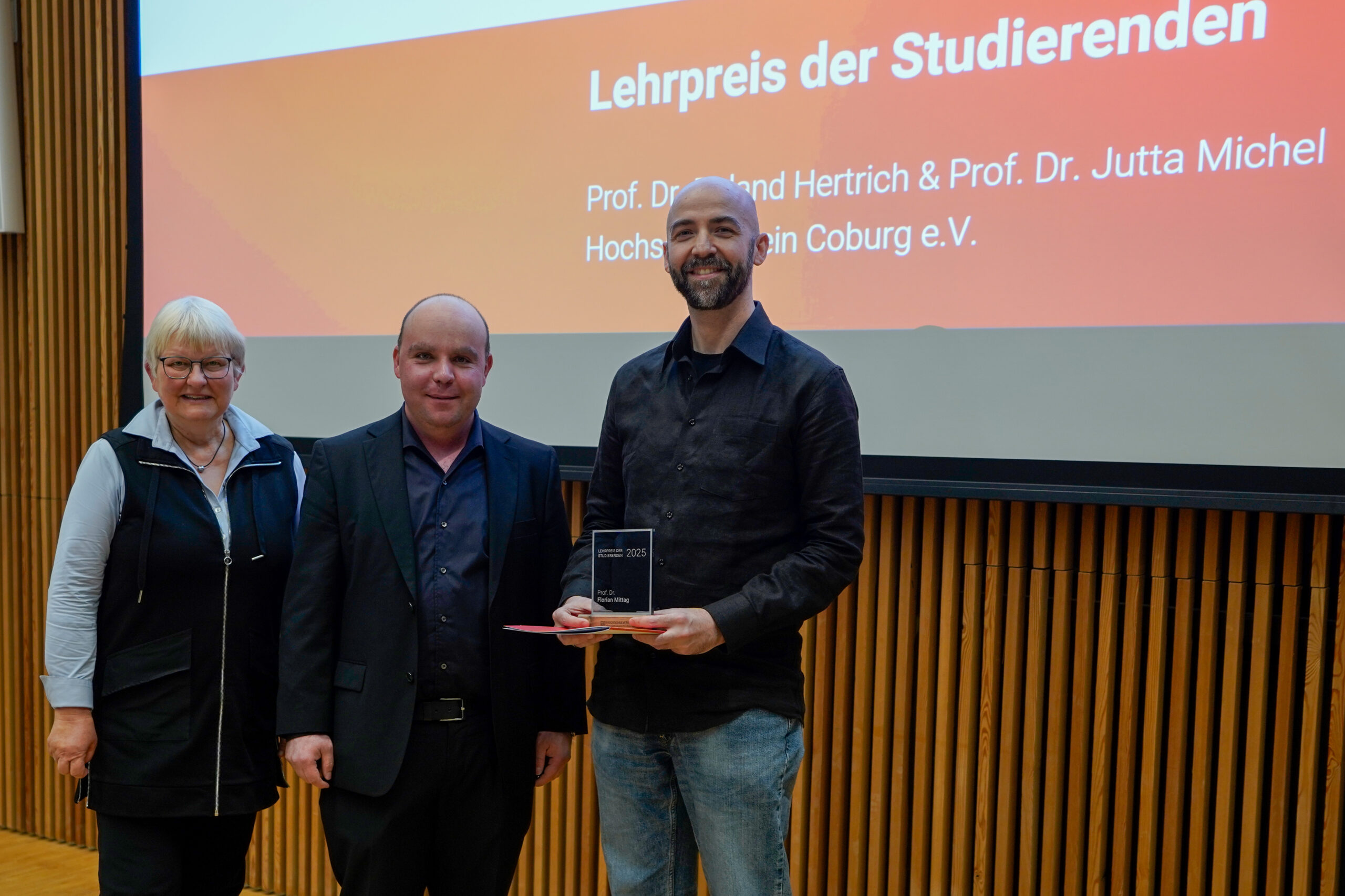Drei Personen stehen in einem Raum vor einem Bildschirm mit der Aufschrift "Lehrpreis der Studierenden". Die Person auf der rechten Seite hält eine Auszeichnung in der Hand. Alle drei lächeln und freuen sich über ihre Anerkennung als beliebteste Lehrende. at Hochschule Coburg
