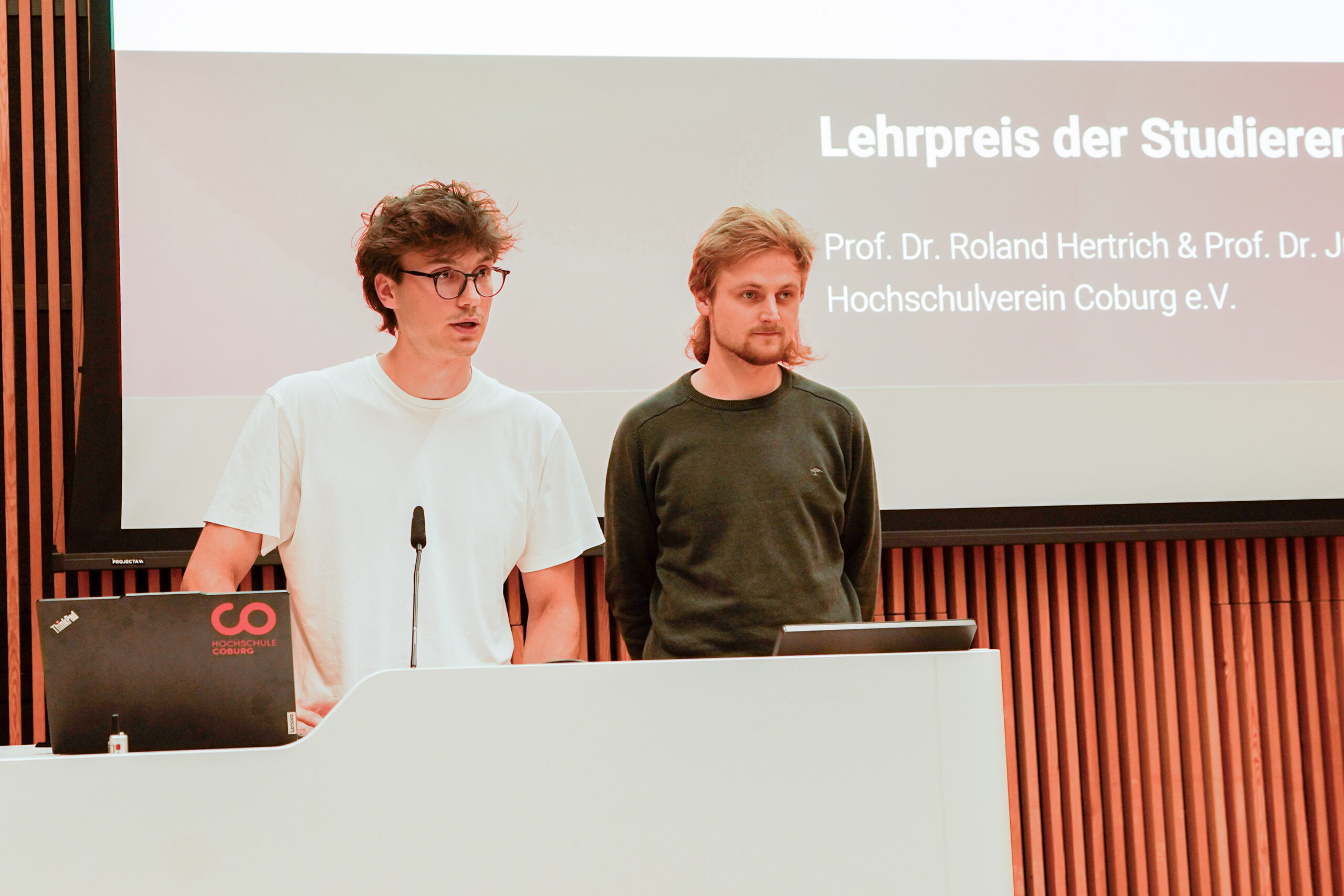 Zwei junge Männer stehen an einem Podium und halten einen Vortrag. Einer trägt ein weißes T-Shirt und eine Brille, der andere einen dunklen Pullover. Auf einem großen Bildschirm hinter ihnen wird ein deutscher Text über die innovativste Lehre und einen Lehrpreis angezeigt. at Hochschule Coburg