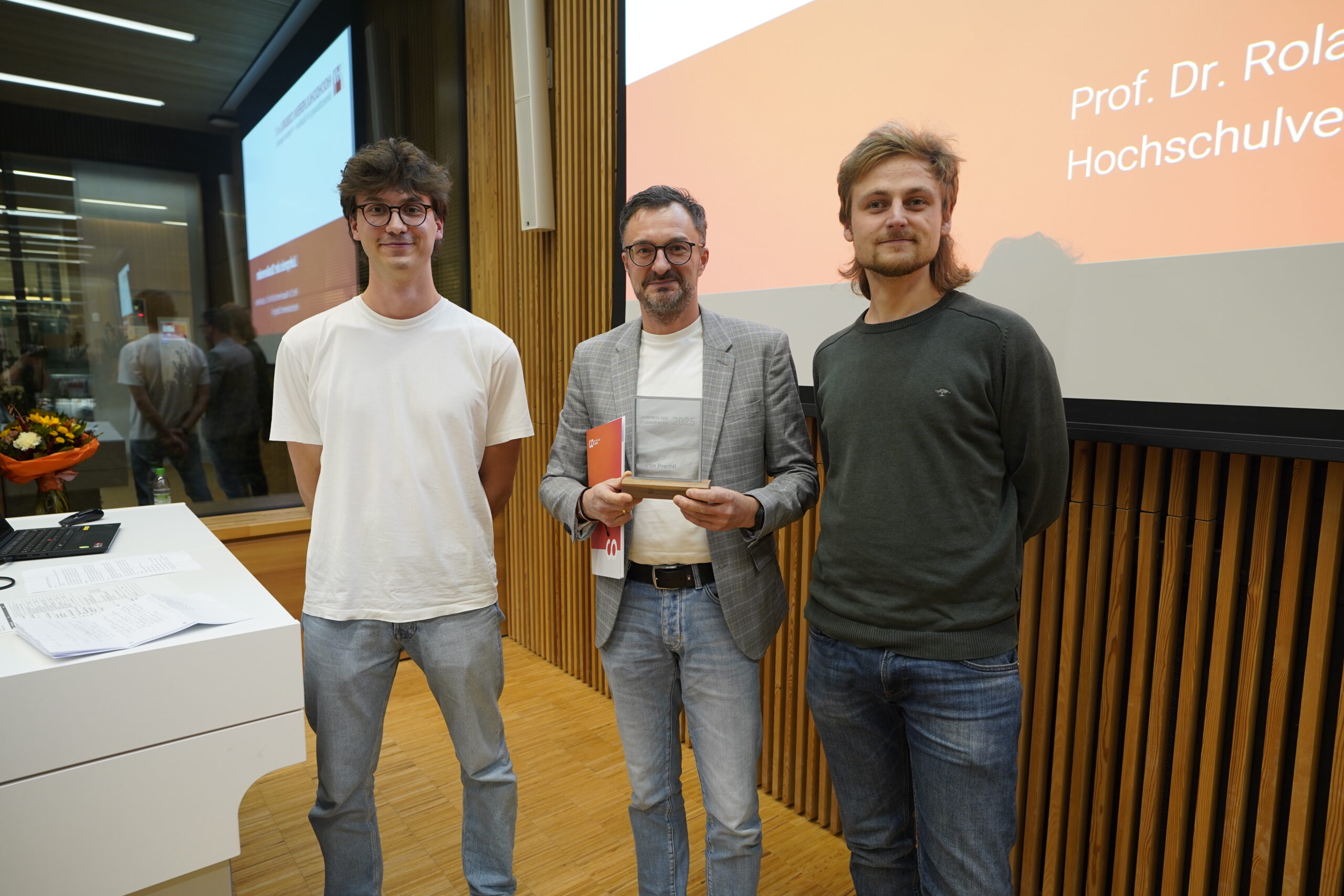 Drei Männer stehen in einem Innenraum vor einem Bildschirm und einer Holztafelwand. Der Mann in der Mitte, ausgezeichnet für innovative Lehre, hält eine Auszeichnung und eine Mappe. Die beiden anderen Lehrenden posieren neben ihm, mit einem Blumenstrauß auf einem nahen weißen Tisch. at Hochschule Coburg