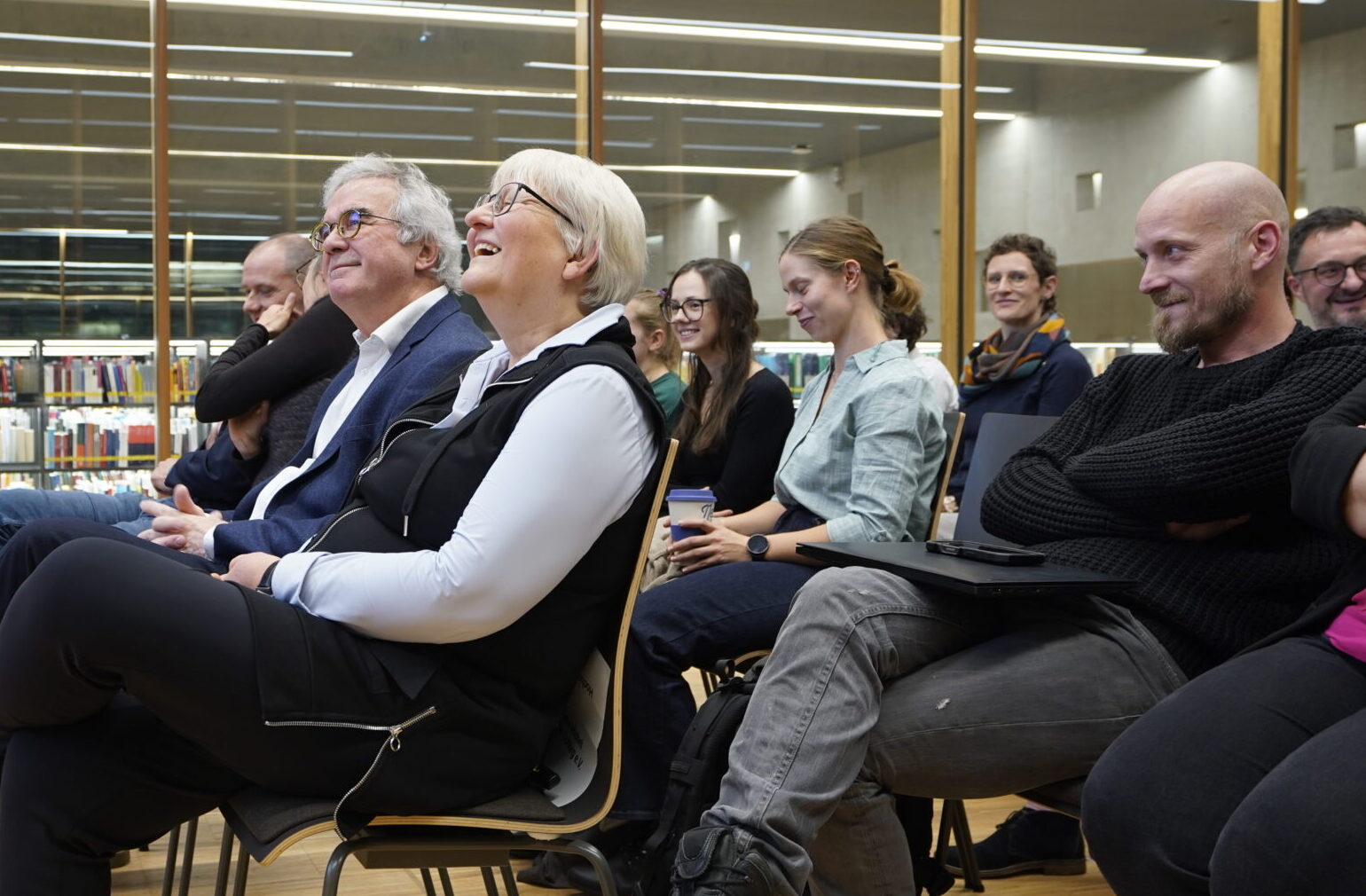 Eine Gruppe von Menschen sitzt auf Stühlen in einer modernen Bibliothek und beobachtet lächelnd und lachend das innovative Lehrende außerhalb des Bildausschnitts. Im Hintergrund sind durch große Fenster Bücherregale zu sehen. at Hochschule Coburg