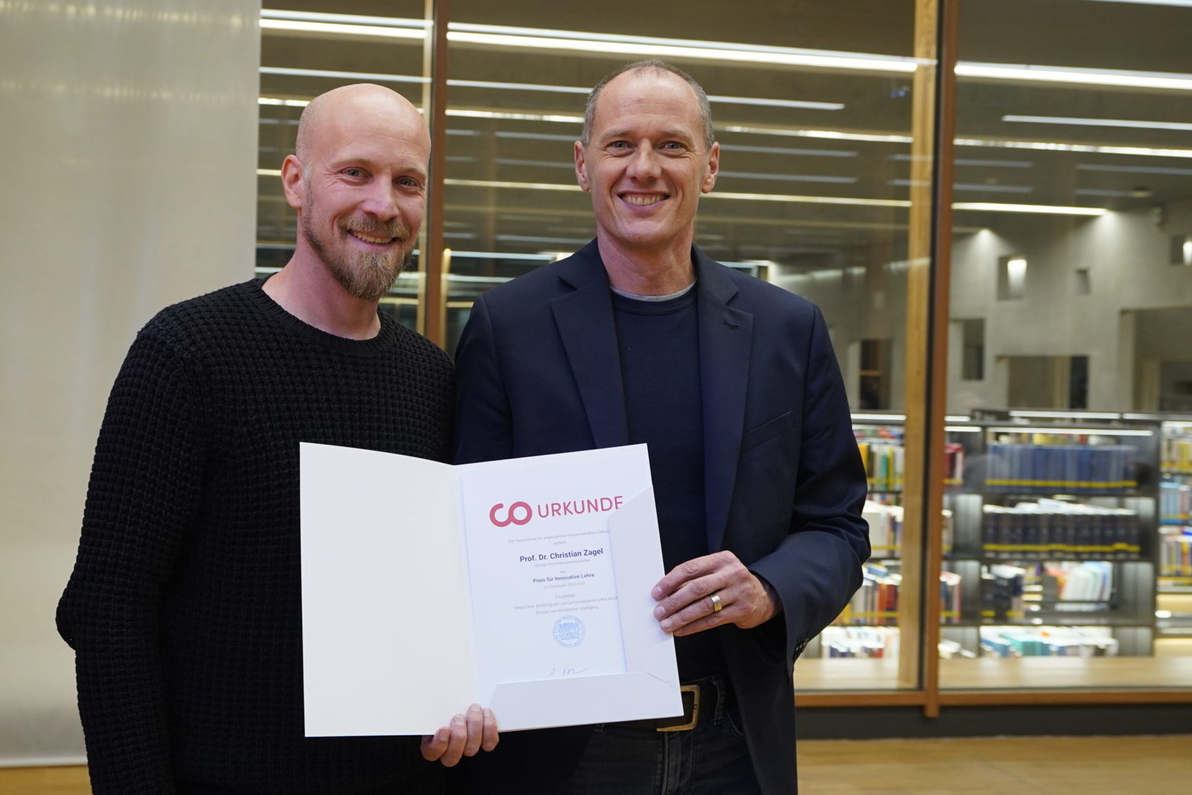 Zwei lächelnde Männer stehen in einem Haus, einer hält eine geöffnete Urkundenmappe mit der Aufschrift "Urkunde". Sie posieren gemeinsam vor großen Fenstern, die innovative Lehre und ihre Anerkennung als beliebte Lehrende feiern, mit Regalen und Büchern im Hintergrund. at Hochschule Coburg