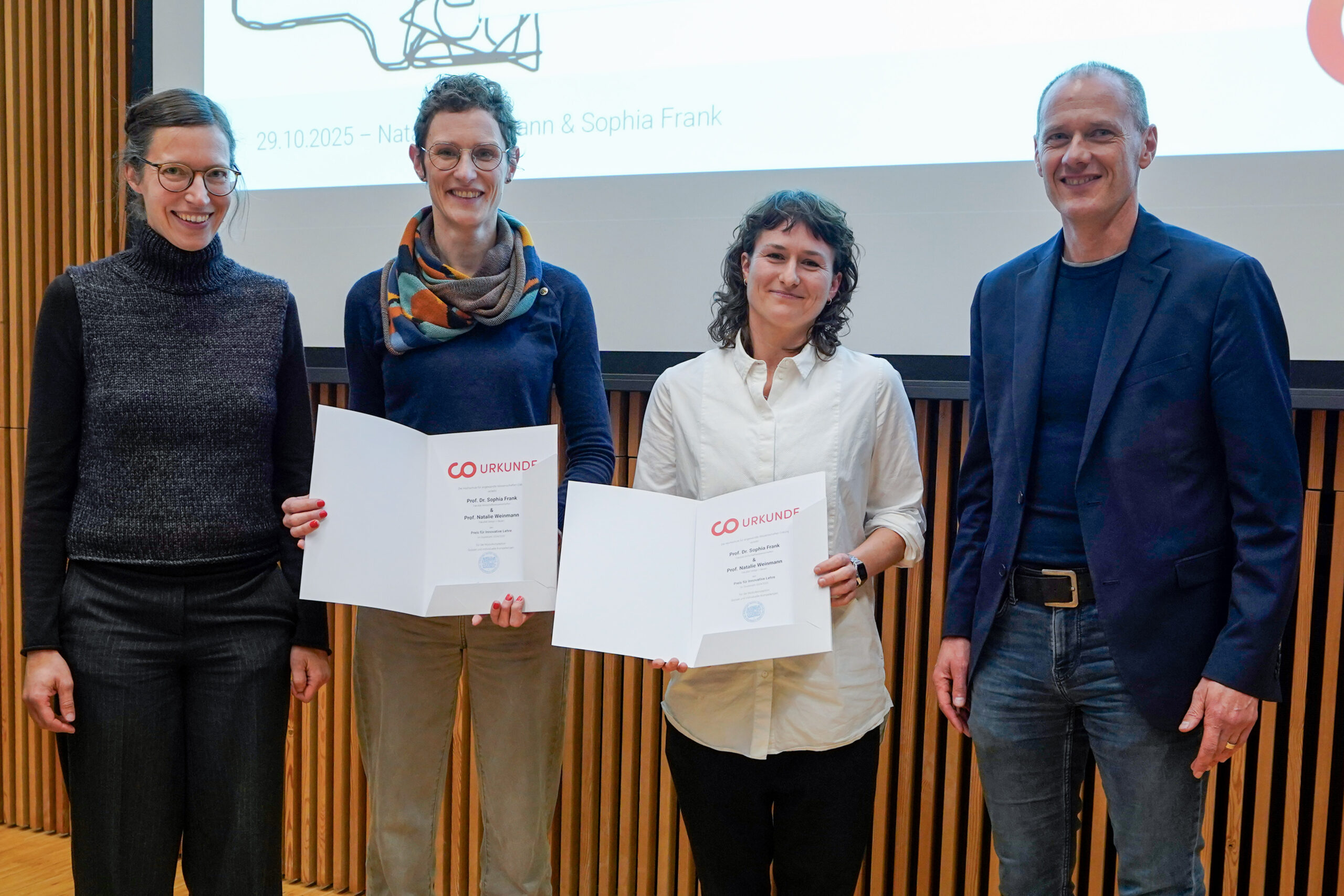 Vier Personen stehen in einem Innenraum vor einem Präsentationsbildschirm und einer Holztafelwand; zwei Frauen in der Mitte, ausgezeichnet als innovative Lehrende, halten Urkunden und lächeln, während eine weitere Frau und ein Mann zu beiden Seiten stehen und ebenfalls lächeln. at Hochschule Coburg