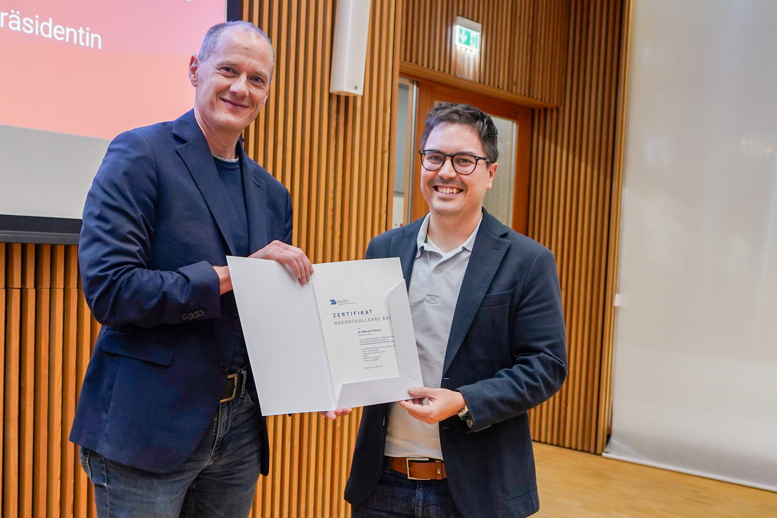 Zwei Männer in Jacken stehen in einem Innenraum, lächeln und halten eine Urkunde während einer Preisverleihung für beliebteste Lehrende in der Hand. Im Hintergrund sind eine Projektionsfläche und holzgetäfelte Wände zu sehen. at Hochschule Coburg