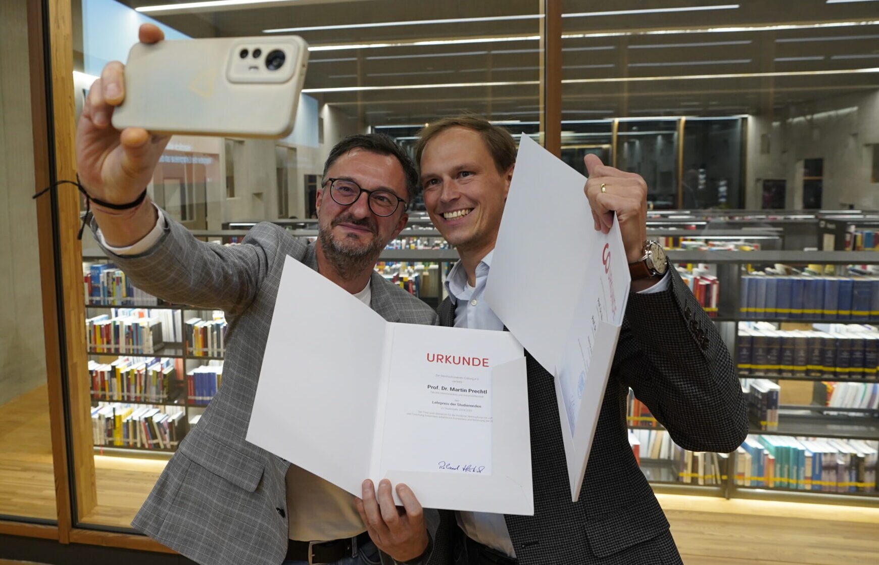 Zwei Männer, die lächelnd und mit Urkunden in der Hand ein Selfie in einer modernen Bibliothek machen, um ihre innovative Lehre zu feiern, mit Regalen voller Bücher im Hintergrund. at Hochschule Coburg
