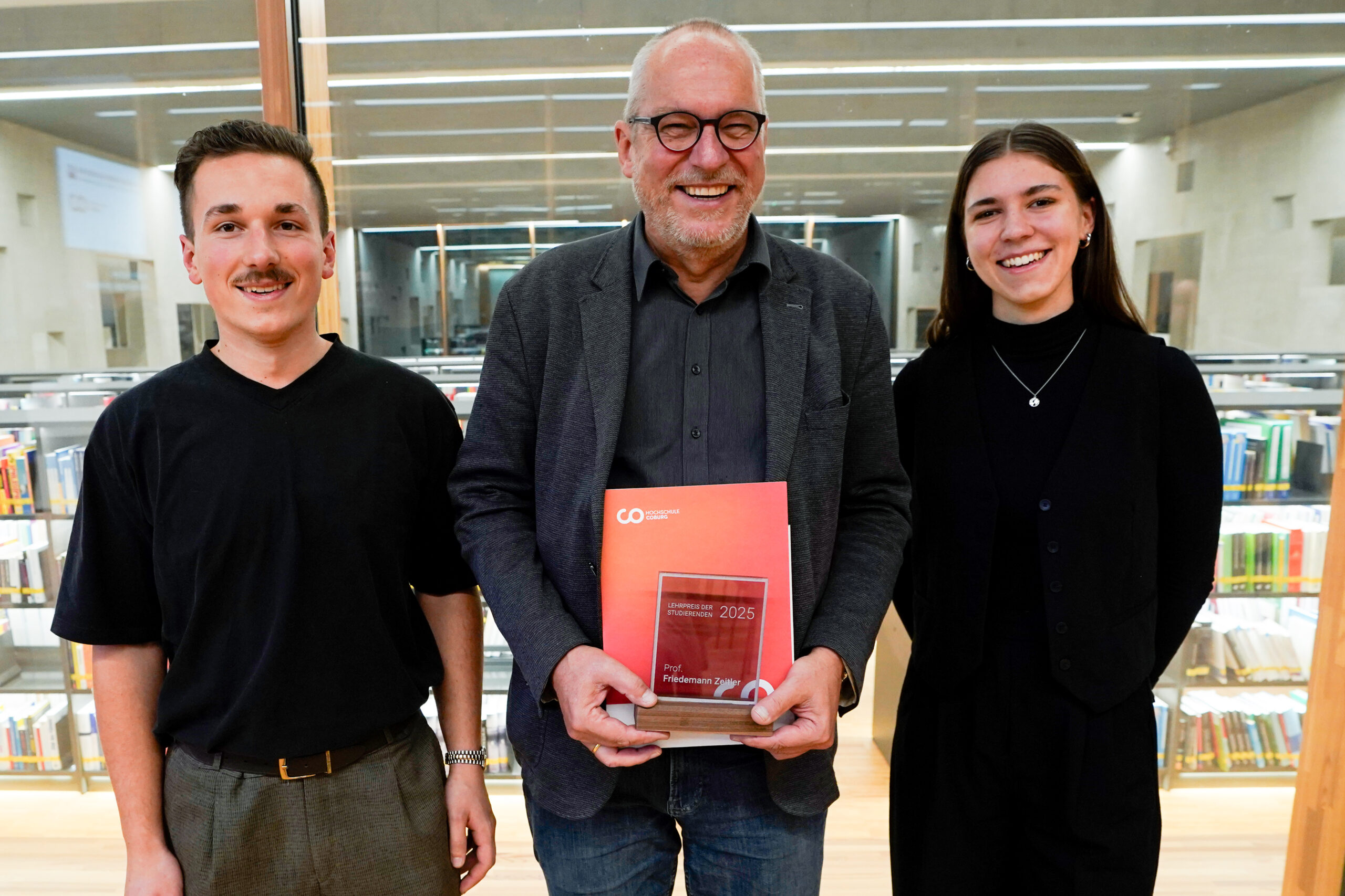 Drei Personen stehen lächelnd in einer Bibliothek. Die Person in der Mitte, die als eine der innovativsten Lehrenden geehrt wurde, hält eine rote Plakette und eine Mappe, flankiert von einem Mann und einer Frau. Bücherregale füllen den Hintergrund. at Hochschule Coburg
