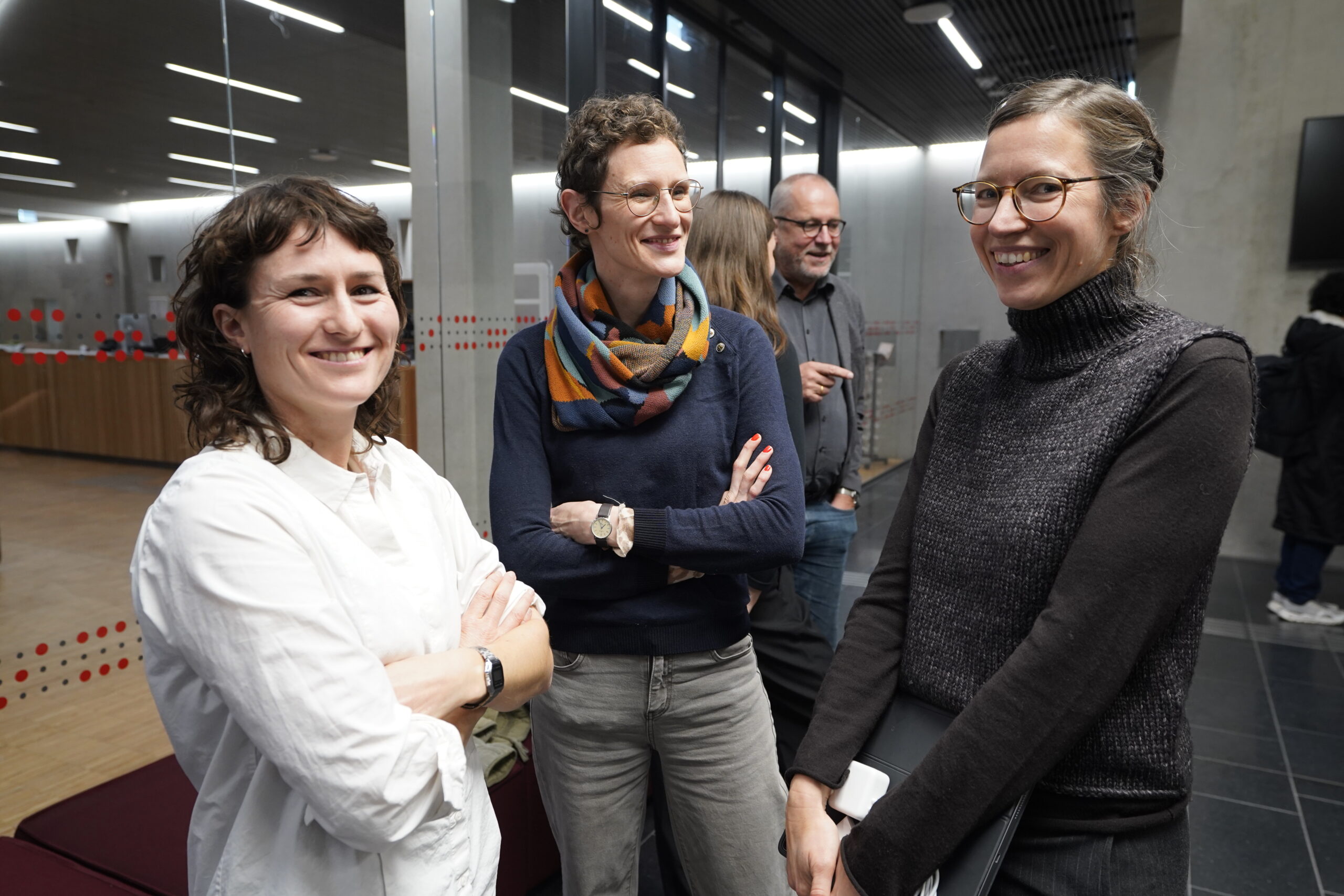 Drei Frauen stehen in einem Innenraum zusammen, lächeln und unterhalten sich - eine in einem weißen Hemd, eine andere mit einem bunten Schal und die dritte in einem grauen Pullover. Zwei Männer unterhalten sich im Hintergrund und spiegeln einen innovativen Geist der Lehre unter engagierten Lehrenden wider. at Hochschule Coburg