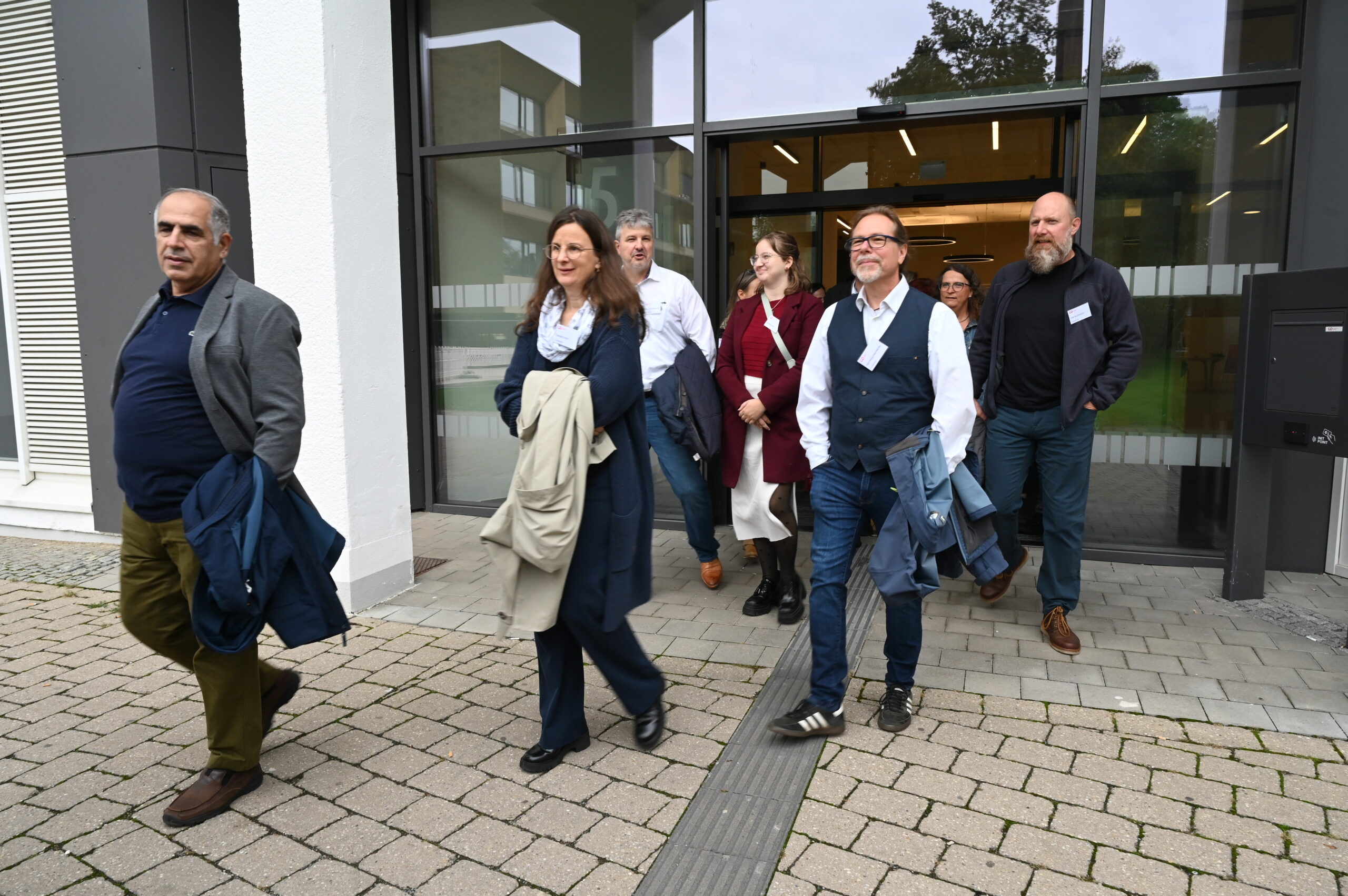 Eine Gruppe von neun Erwachsenen, möglicherweise alte Freunde von der Hochschule, gekleidet in Geschäfts- oder Freizeitkleidung, tritt aus einem modernen Gebäude mit Glastüren und scheint sich zu unterhalten und zu lächeln. at Hochschule Coburg