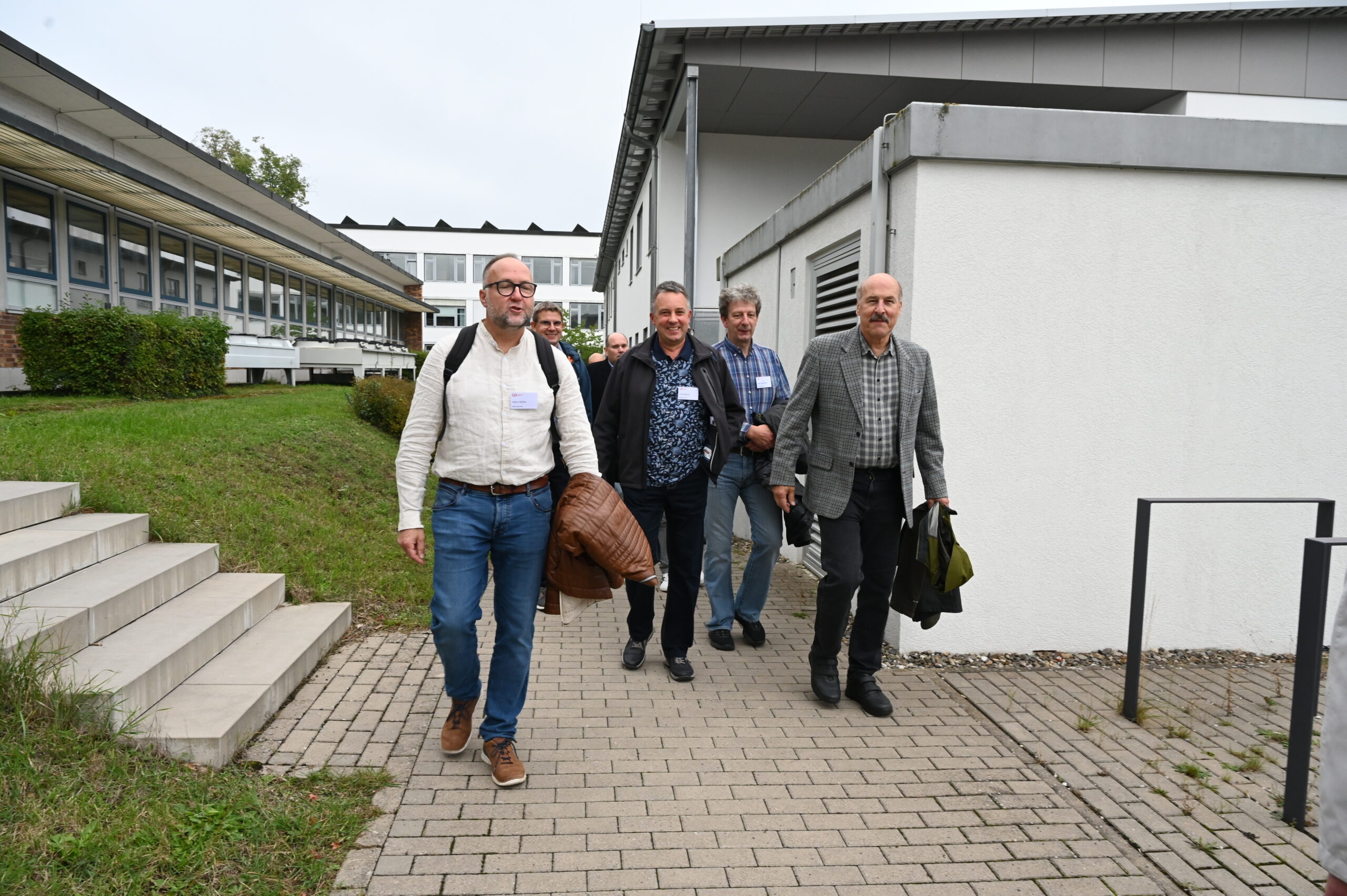 Vier Männer, möglicherweise alte Freunde von der Hochschule, gehen mit Taschen und Jacken einen gepflasterten Weg entlang, der an modernen Gebäuden vorbeiführt. Sie lächeln und sind leger oder halb-formal gekleidet, vielleicht nehmen sie an einem Silberjubiläum oder einer besonderen Veranstaltung teil. at Hochschule Coburg