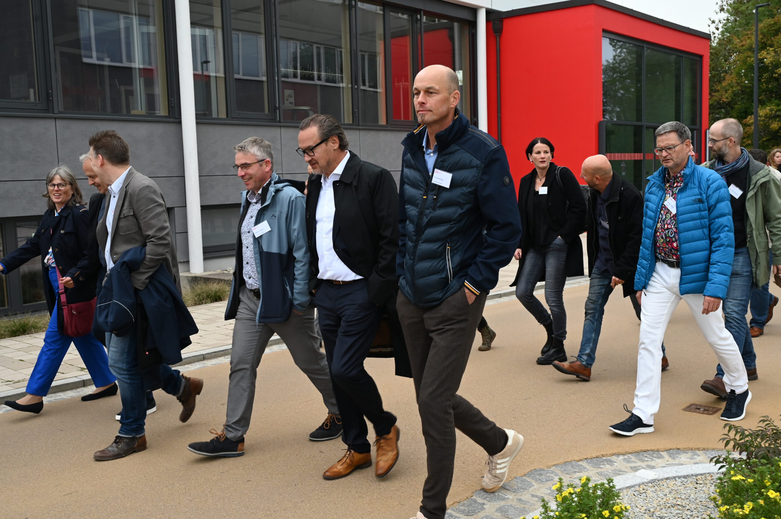Eine Gruppe professionell gekleideter Erwachsener geht vor einem modernen Hochschulgebäude mit grauen und roten Wänden spazieren, einige tragen Jacken und Namensschilder und unterhalten sich über das bevorstehende Silberjubiläum. at Hochschule Coburg