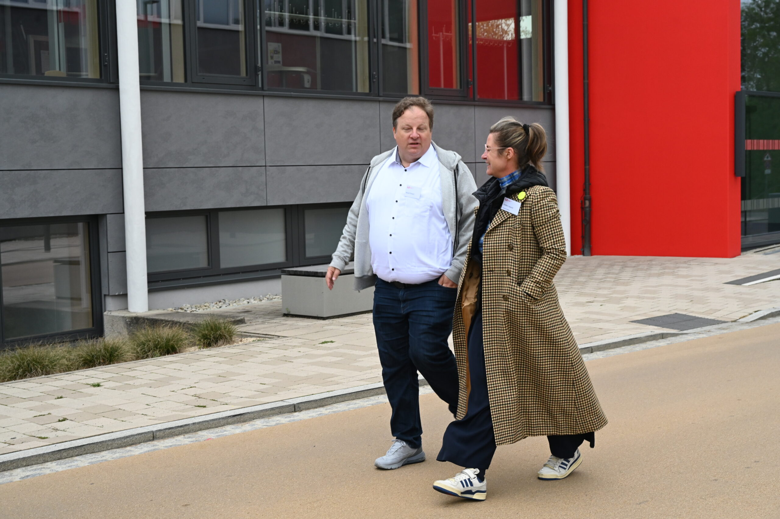 Zwei alte Freunde gehen nebeneinander auf einem gepflasterten Gehweg in der Nähe eines modernen Gebäudes mit großen Fenstern und einem kräftigen roten Akzent. Der Mann trägt ein weißes Hemd und eine helle Jacke, die Frau einen langen, braun karierten Mantel. at Hochschule Coburg