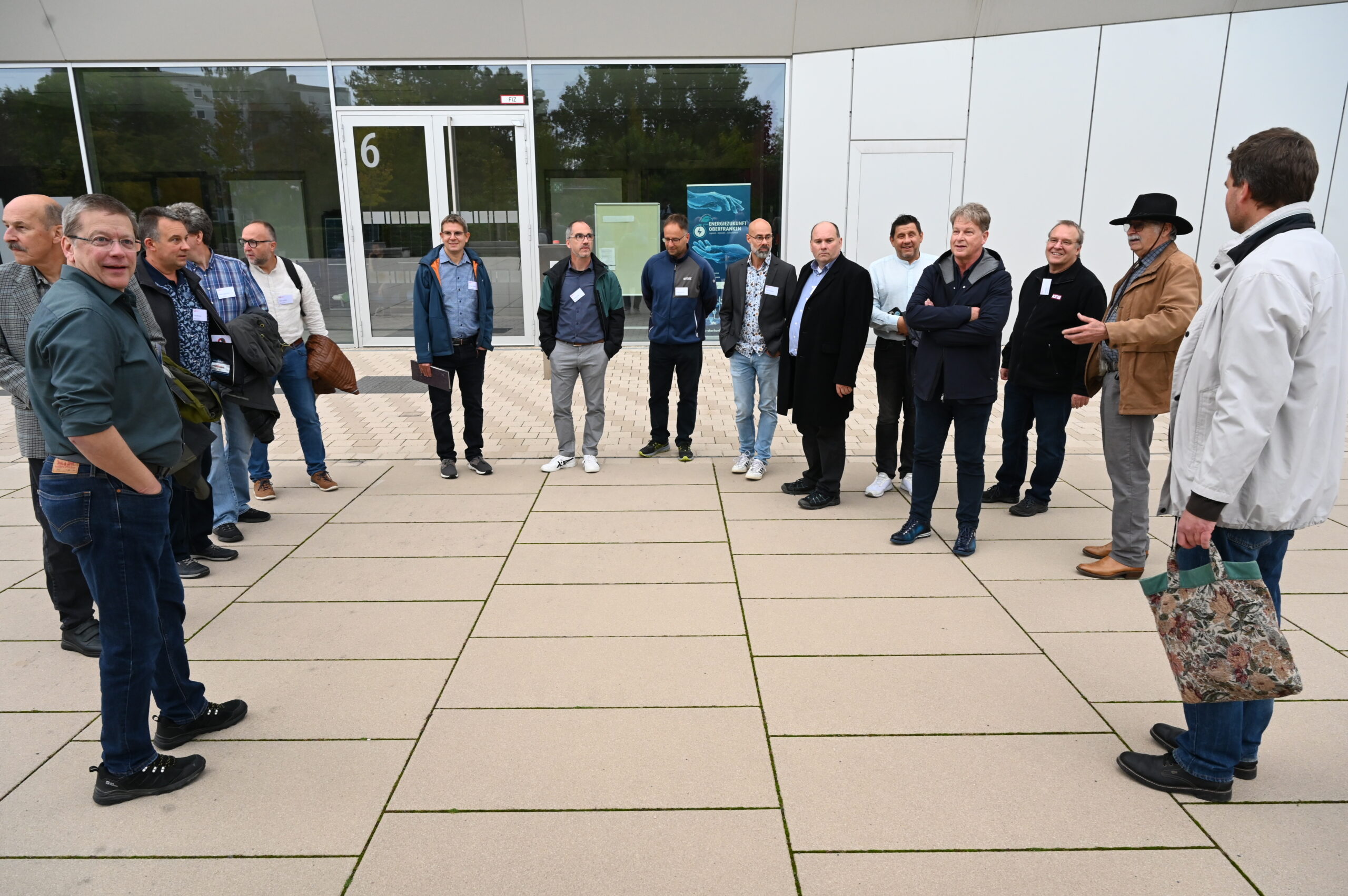 Eine Gruppe von Menschen, wahrscheinlich einige alte Freunde, steht im Halbkreis vor einem modernen Hochschulgebäude mit großen Fenstern und weißen Tafeln. Sie plaudern und hören zu - einige tragen Namensschilder, und einer hält eine gemusterte Tasche, mit der er ein Silberjubiläum feiert. at Hochschule Coburg
