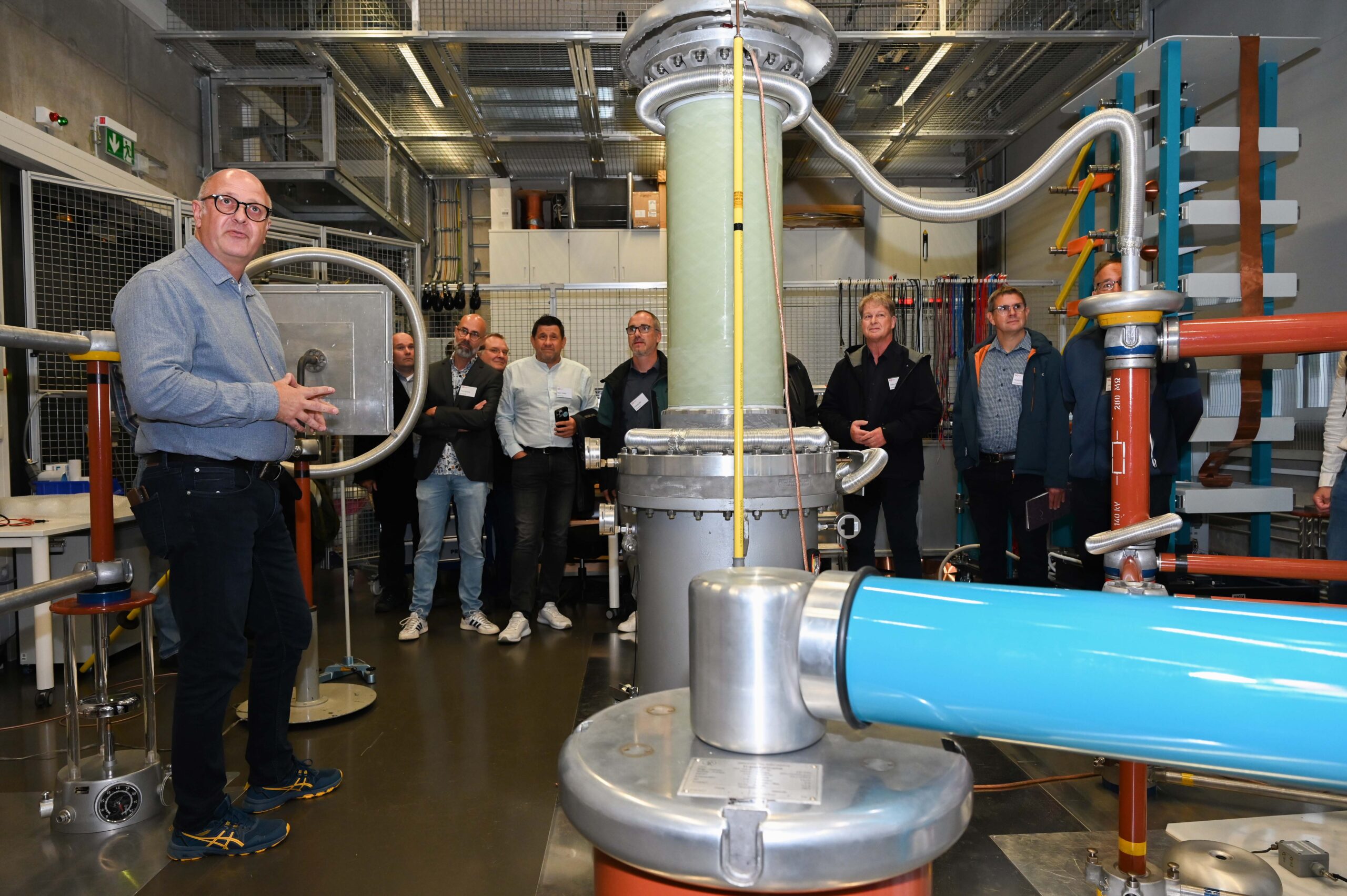 Eine Gruppe von Personen, darunter alte Freunde von der Hochschule, steht in einem Industrielabor und beobachtet einen Mann, der große Metall- und Kunststoffmaschinen mit Schläuchen und Drähten in einer modernen, technischen Umgebung präsentiert. at Hochschule Coburg