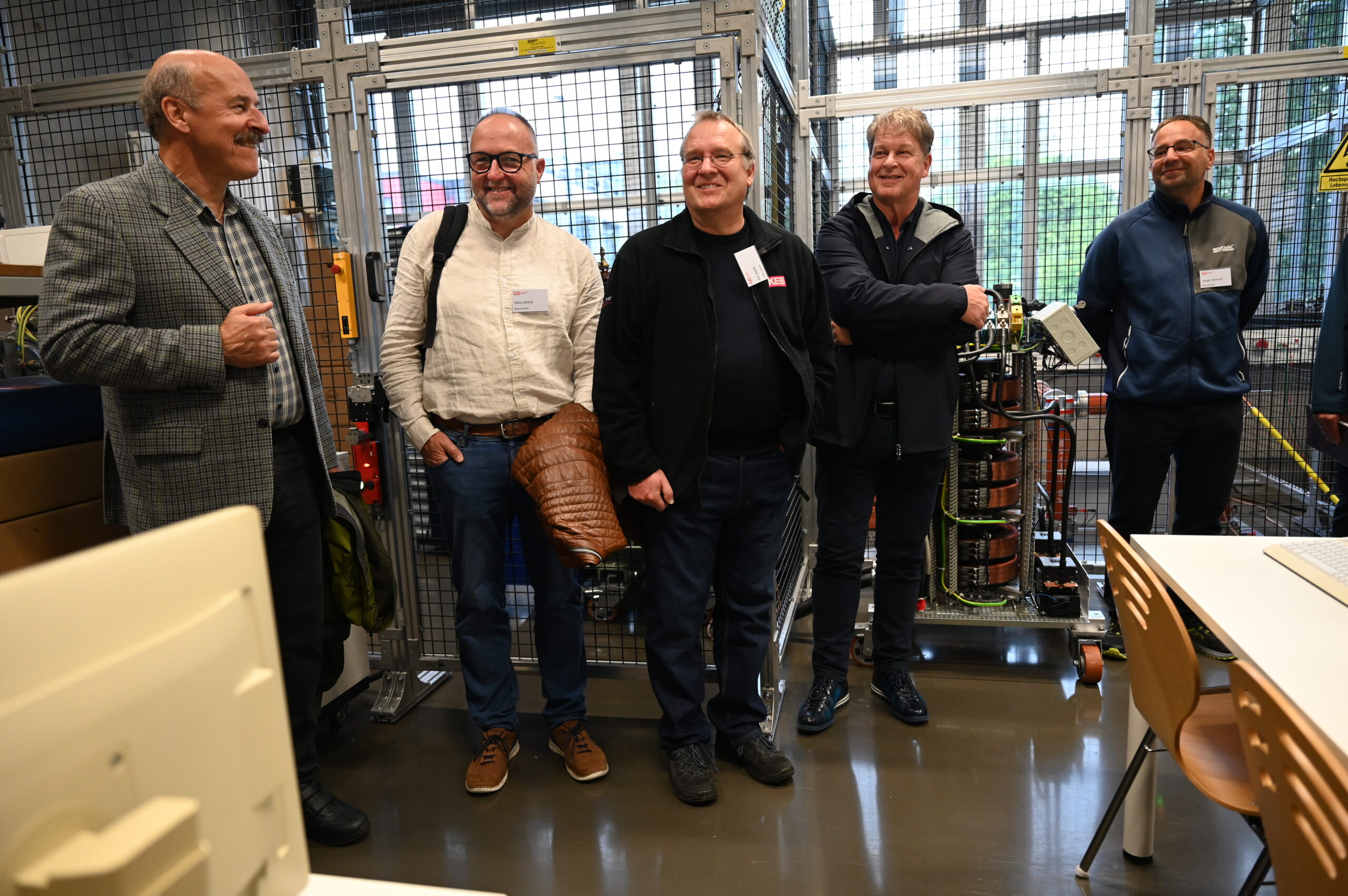 Fünf Männer stehen lächelnd in einer Werkstatt oder einem Labor der Hochschule, mit industriellen Geräten und Metallzäunen im Hintergrund. Sie scheinen alte Freunde zu sein, die sich während einer Besichtigung, vielleicht zu Ehren eines Silberjubiläums, unterhalten. at Hochschule Coburg