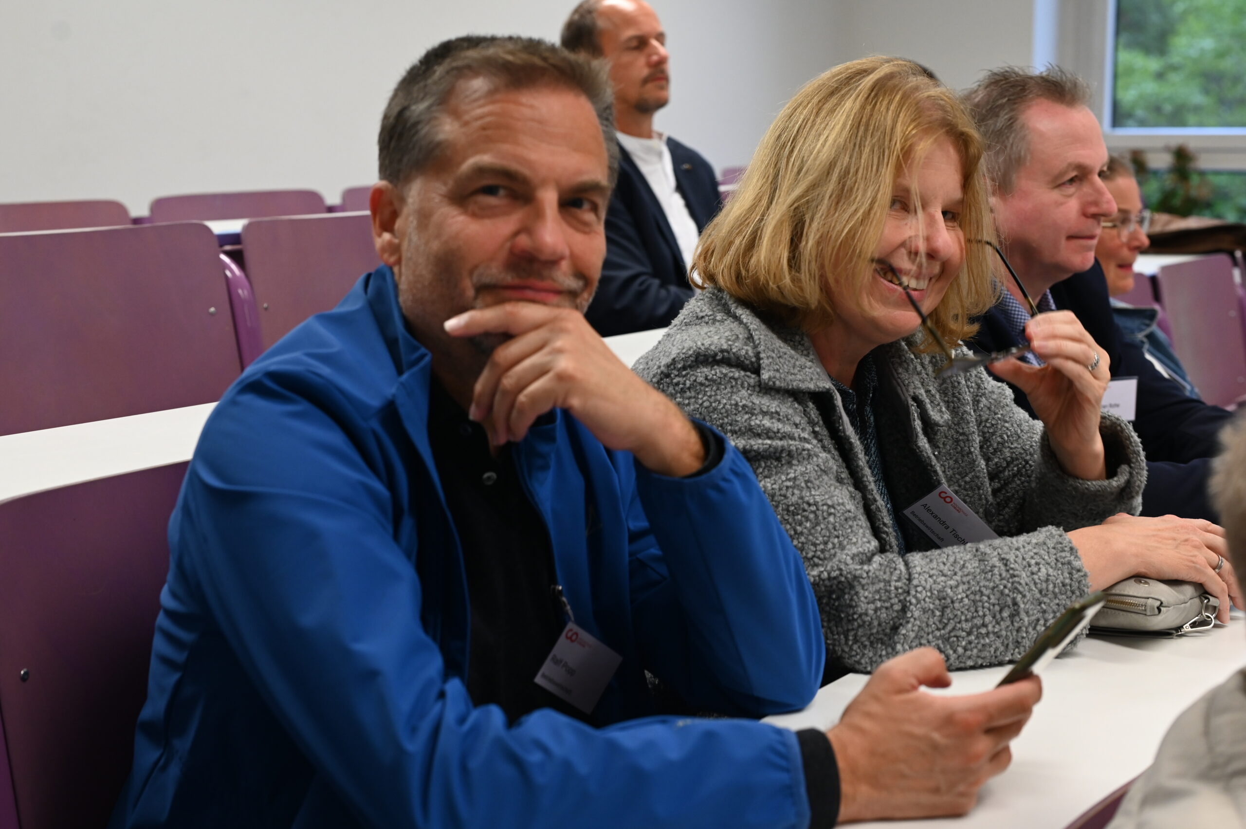 Ein Mann in einer blauen Jacke und eine Frau in einem grauen Pullover lächeln, während sie in einem lila bestuhlten Hörsaal des Silberjubiläums der Hochschule sitzen, umgeben von anderen Zuhörern. Der Mann hält ein Telefon in der Hand und blickt in die Kamera. at Hochschule Coburg