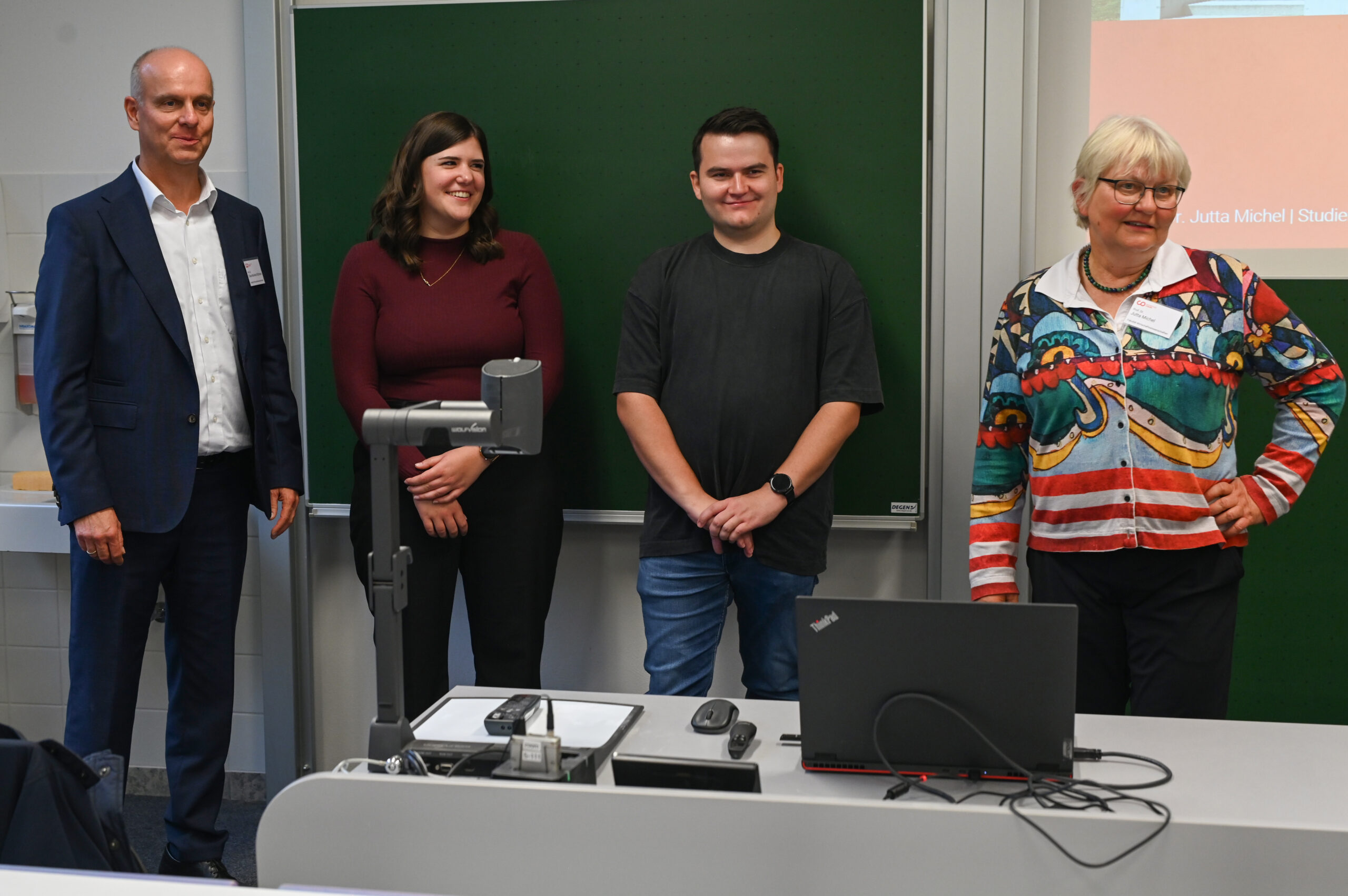 Vier Erwachsene stehen vor einem Klassenzimmer an der Hochschule, drei von ihnen lächeln freundlich. Im Vordergrund steht ein Schreibtisch mit einem Laptop und Geräten, und eine grüne Kreidetafel markiert den Hintergrund - vielleicht um ein besonderes Silberjubiläum zu feiern. at Hochschule Coburg