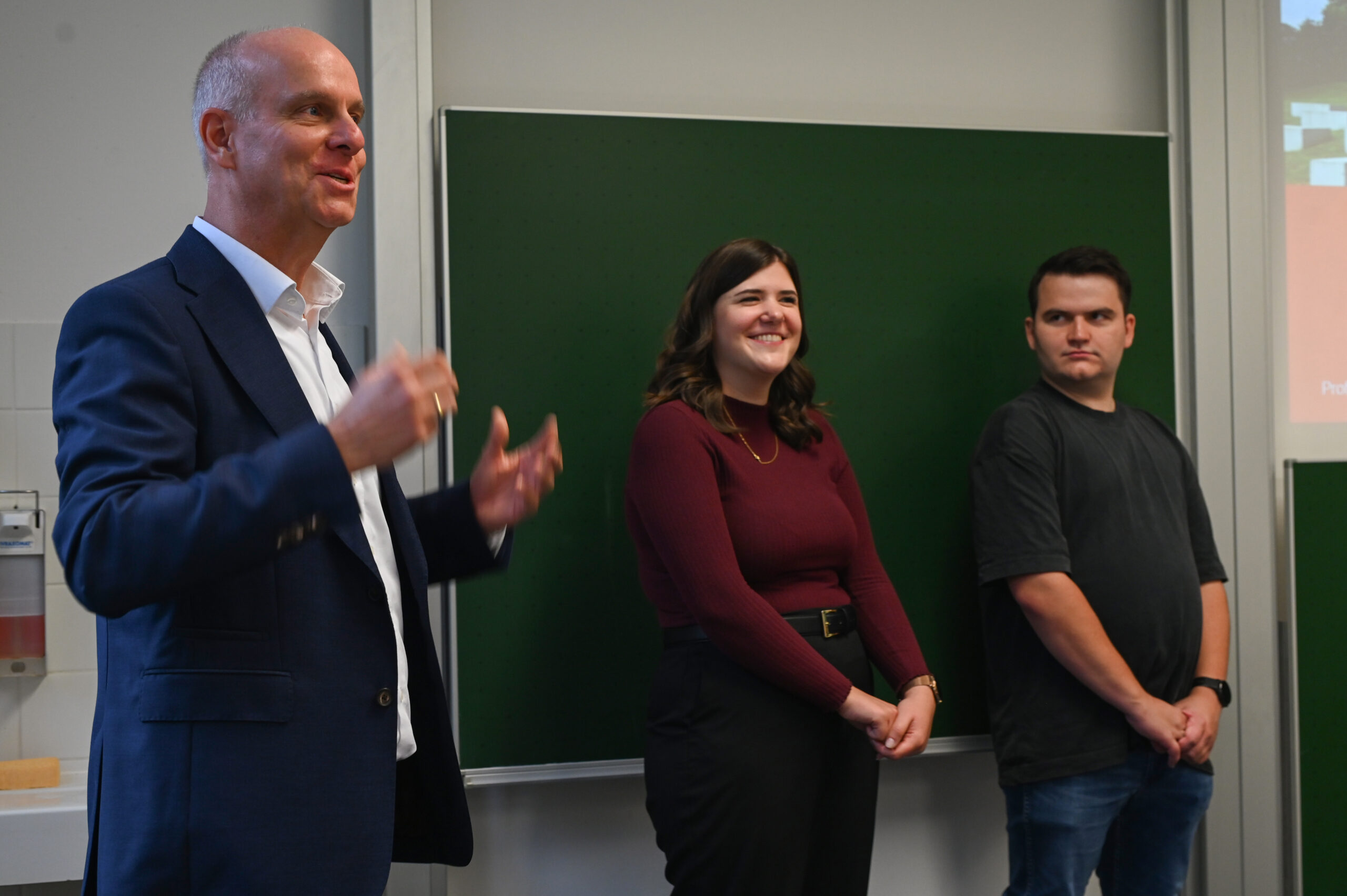 Ein Mann im Anzug spricht angeregt neben zwei jungen Erwachsenen, einer lächelnden Frau in einem kastanienbraunen Oberteil und einem ernsten Mann in Schwarz, vor einer grünen Kreidetafel in der Hochschule, die möglicherweise ihr Silberjubiläum feierte. at Hochschule Coburg