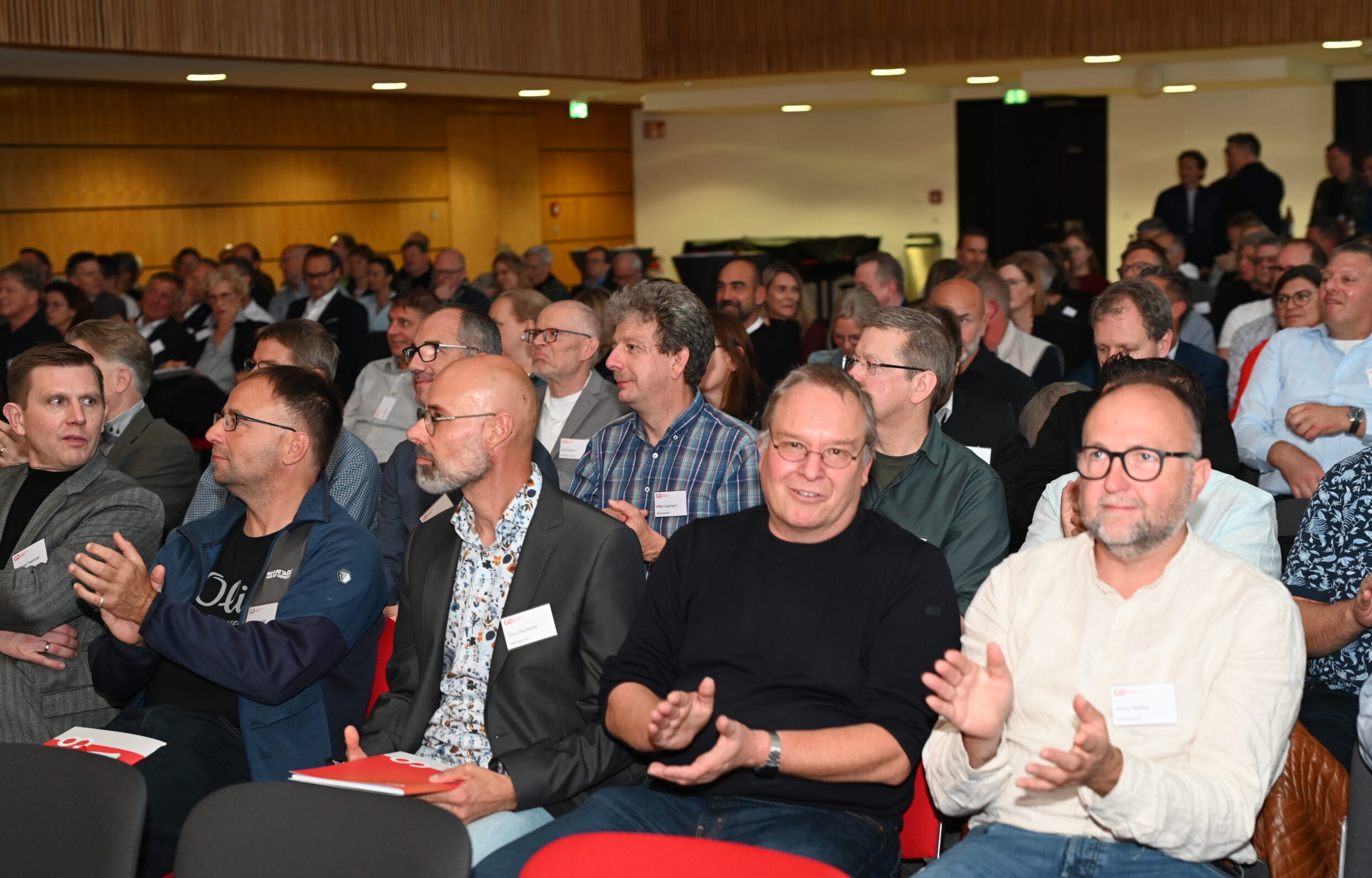 Eine große Gruppe von Menschen, meist Männer, sitzt in Stuhlreihen bei einer Indoor-Veranstaltung zur Feier des Silberjubiläums einer Hochschule. Einige klatschen und lächeln alten Freunden zu, während andere in dem gut beleuchteten Raum mit einer Holzwand im Hintergrund aufmerksam zuhören. at Hochschule Coburg