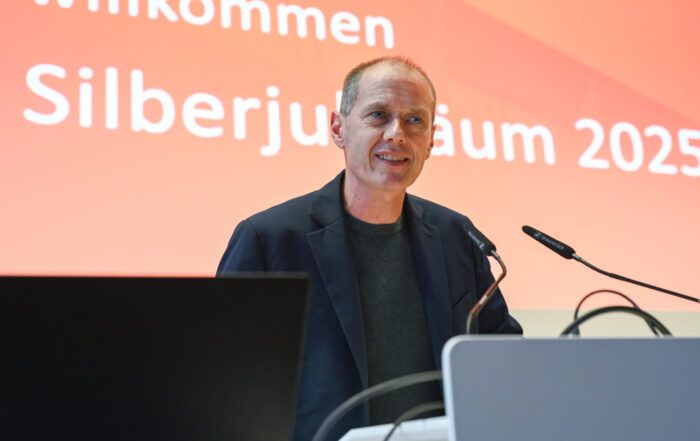 Ein Mann in einer dunklen Jacke steht an einem Podium und spricht in Mikrofone, während hinter ihm auf rotem Hintergrund ein Willkommensgruß und "Silberjubiläum 2025" der Hochschule zu lesen ist. at Hochschule Coburg