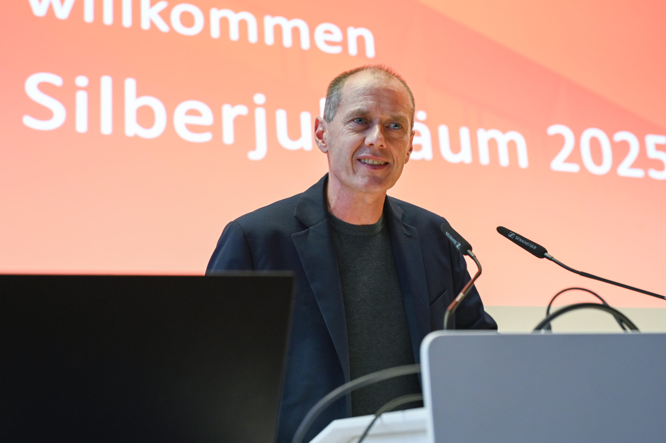 Ein Mann in einer dunklen Jacke steht an einem Podium und spricht in Mikrofone, während hinter ihm auf rotem Hintergrund ein Willkommensgruß und "Silberjubiläum 2025" der Hochschule zu lesen ist. at Hochschule Coburg