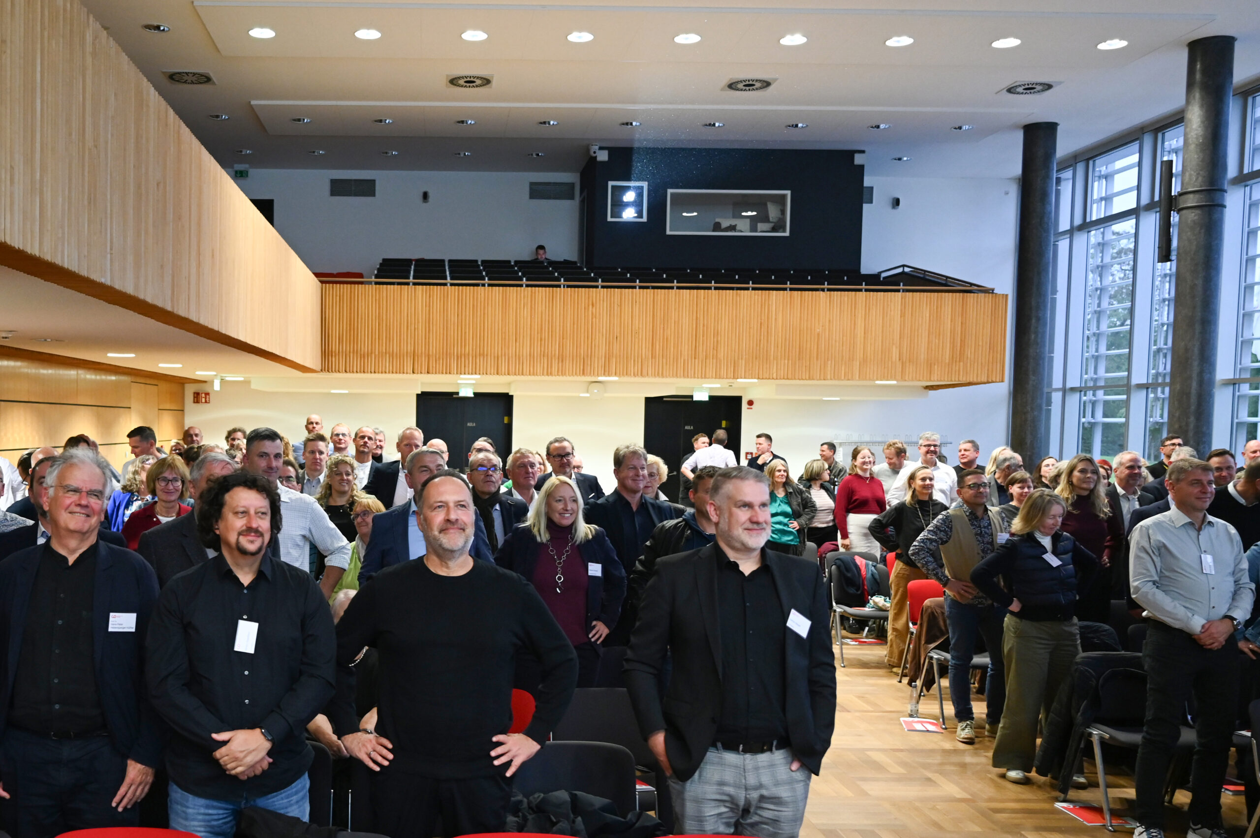 Eine große Gruppe von Menschen, darunter alte Freunde, steht lächelnd in einem gut beleuchteten Konferenzraum mit Holzakzenten und großen Fenstern im Silberjubiläum der Hochschule. Einige halten Papiere in der Hand, im Vordergrund sind leere Stühle zu sehen. at Hochschule Coburg