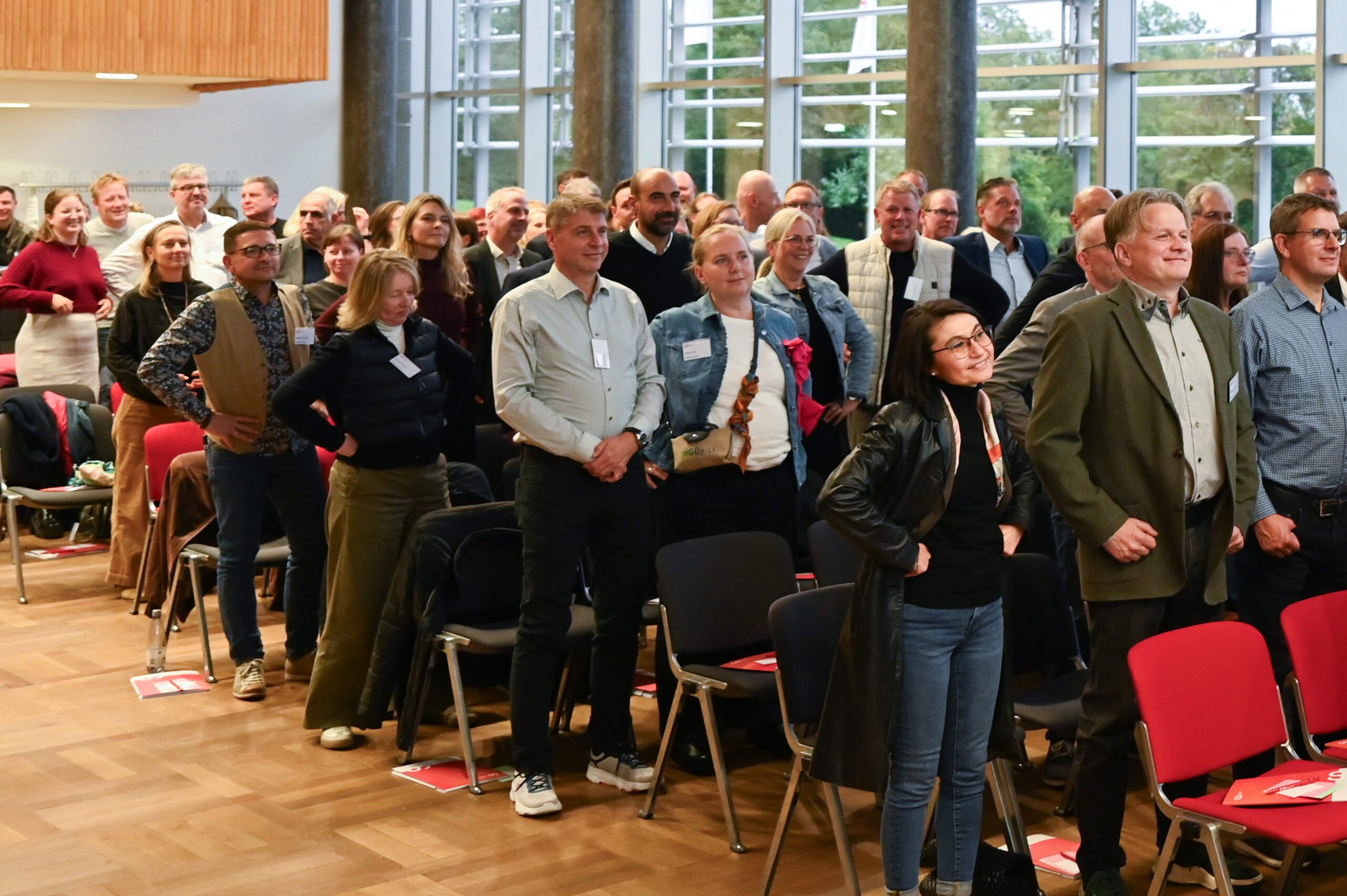 In einem hell erleuchteten Raum mit großen Fenstern steht eine Gruppe von Menschen und lächelt, die an einem Seminar oder einer Konferenz der Hochschule teilnehmen. Einige Stühle sind besetzt, andere leer, und auf mehreren Plätzen liegen rote Mappen zum Silberjubiläum. at Hochschule Coburg
