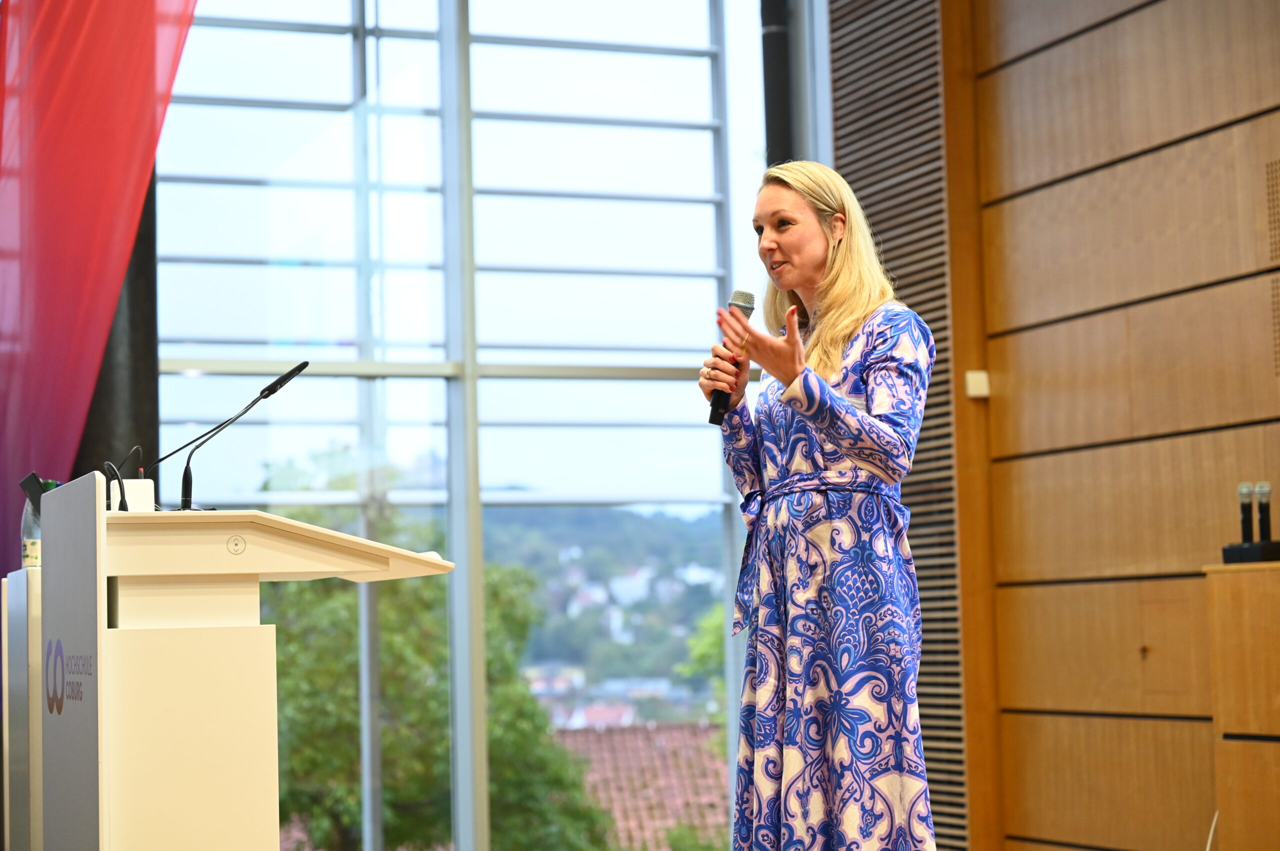 Eine Frau in einem blau-weiß gemusterten Kleid spricht in ein Mikrofon, während sie auf einer Bühne im modernen Konferenzraum der Hochschule steht, die ihr Silberjubiläum inmitten großer Fenster und holzgetäfelter Wände feiert. at Hochschule Coburg